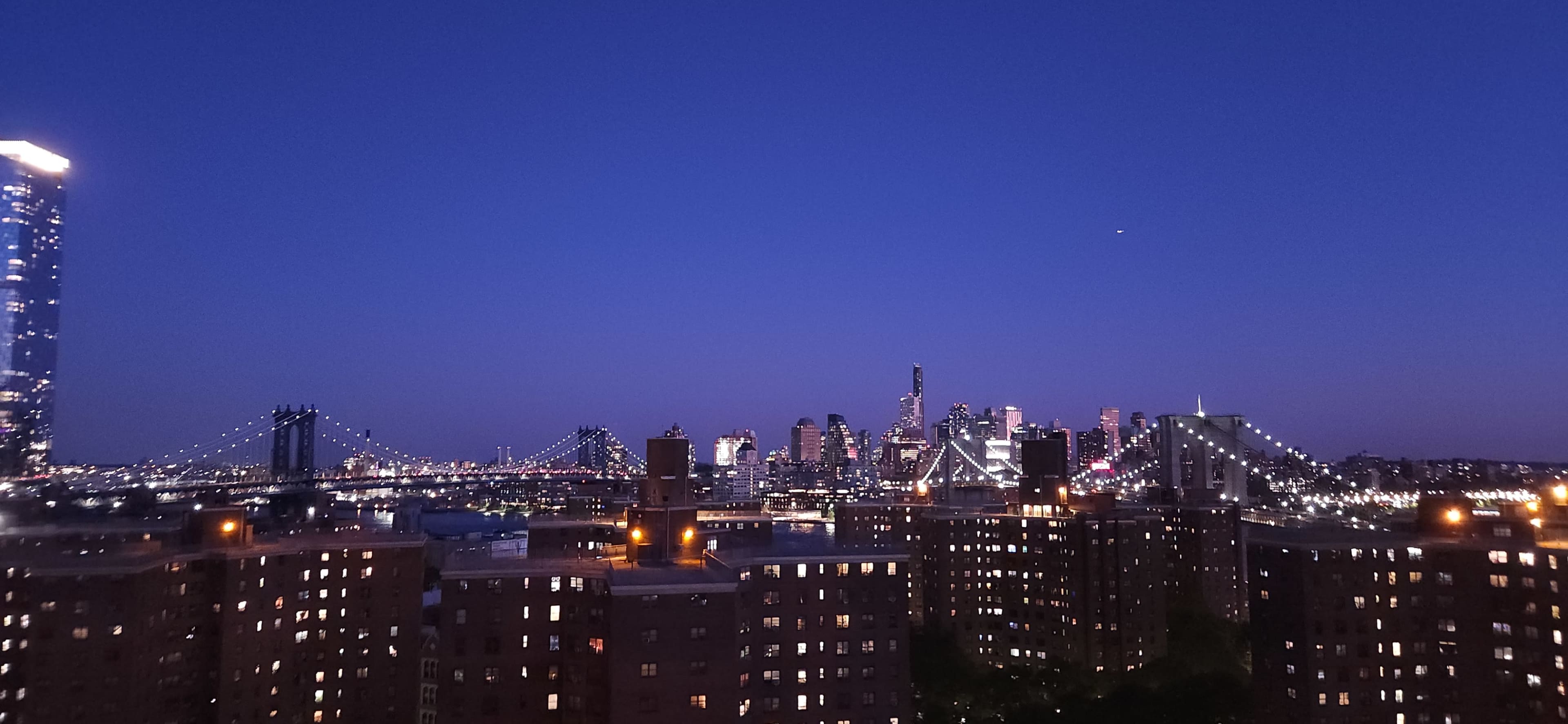 The skyline of a city is illuminated at dusk, featuring bridges and tall buildings against a deep blue sky.