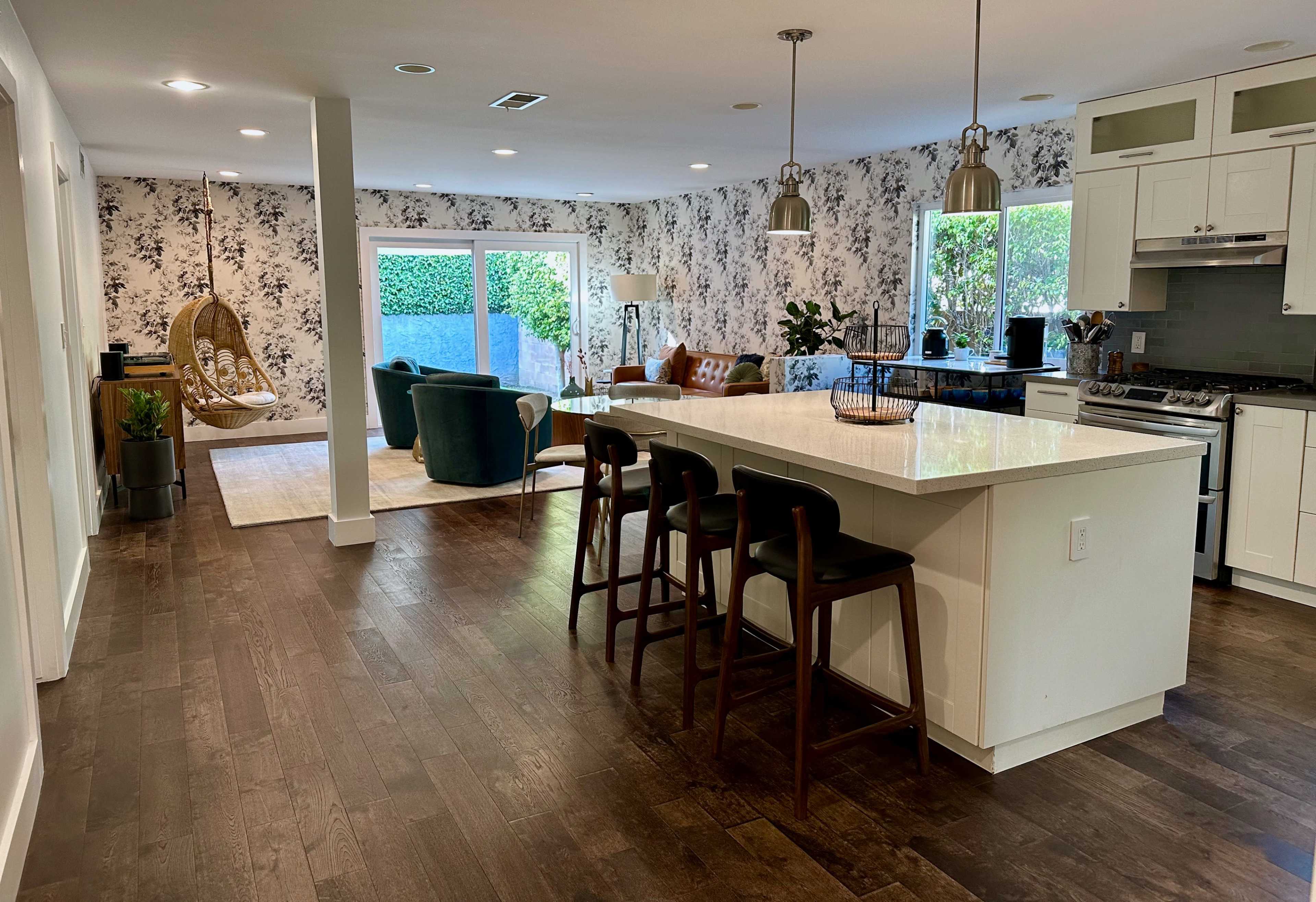 The image shows a modern open-concept living space with a kitchen bar, a dining area, and a cozy lounge area featuring a hanging chair and floral wallpaper.