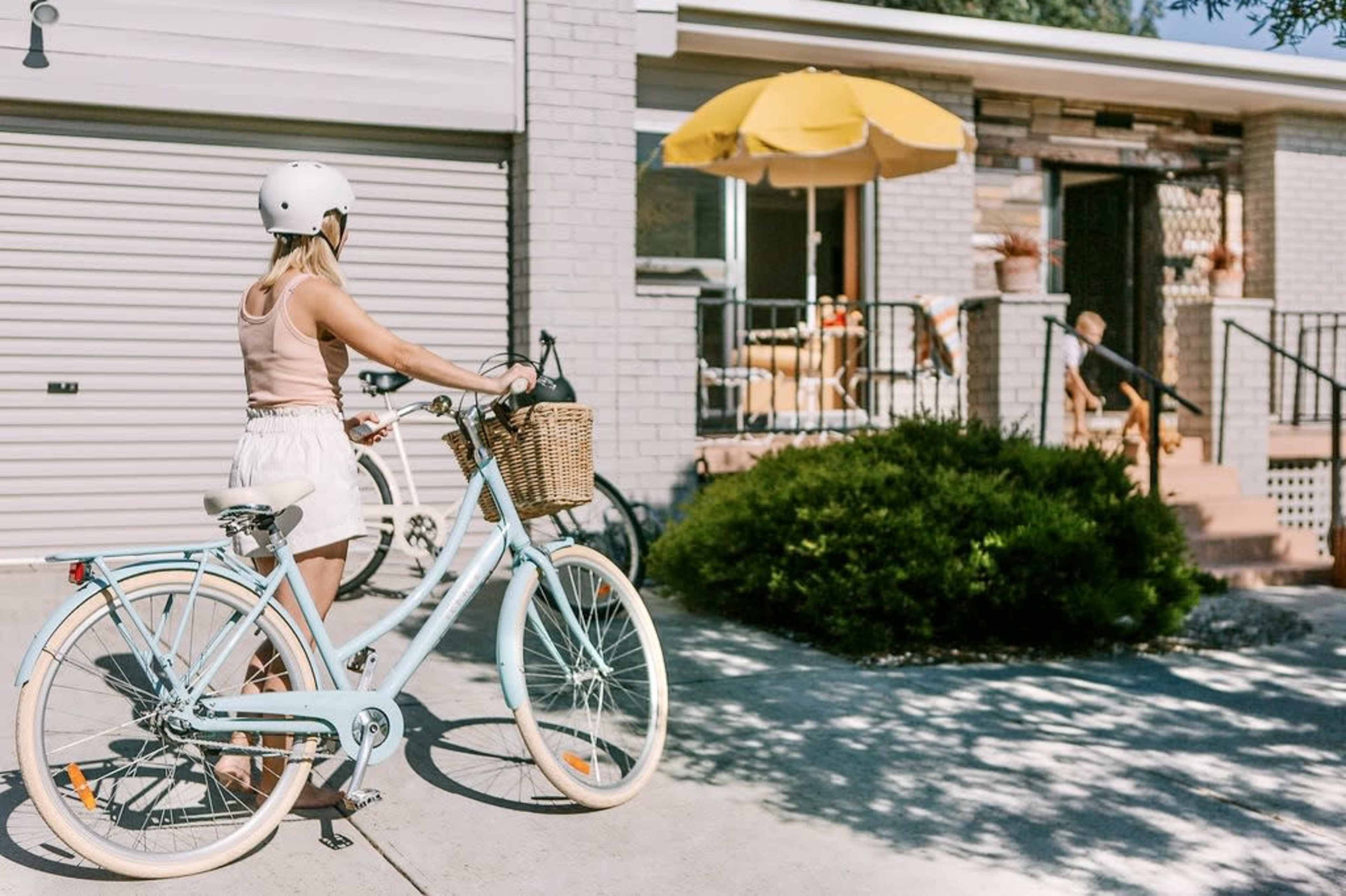 A woman in a helmet pushes a light blue bicycle with a wicker basket along a driveway in front of a house.