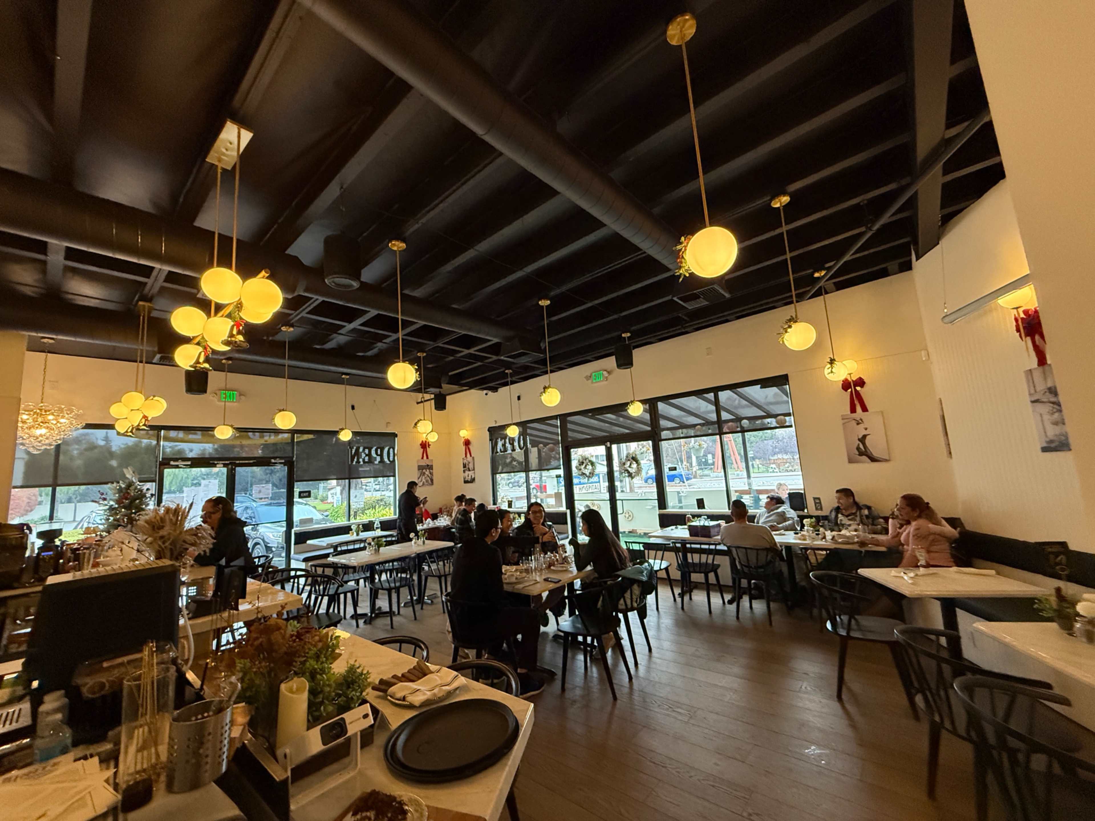 The image shows the interior of a restaurant with black wooden ceiling beams, round tables, and large windows allowing natural light to enter, while patrons are seated at various tables.
