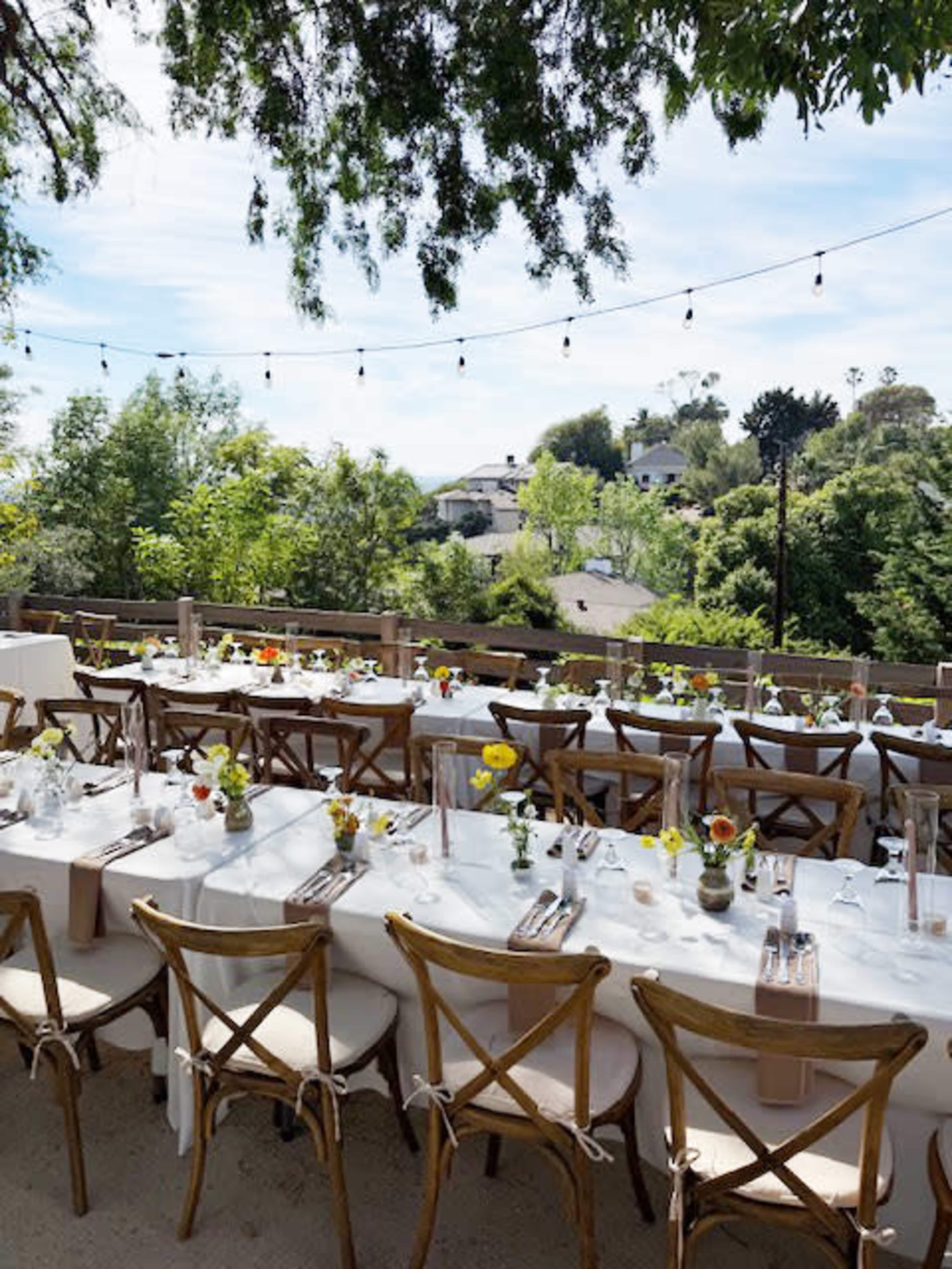 A long dining table is set outside under string lights, surrounded by greenery and with houses visible in the background.