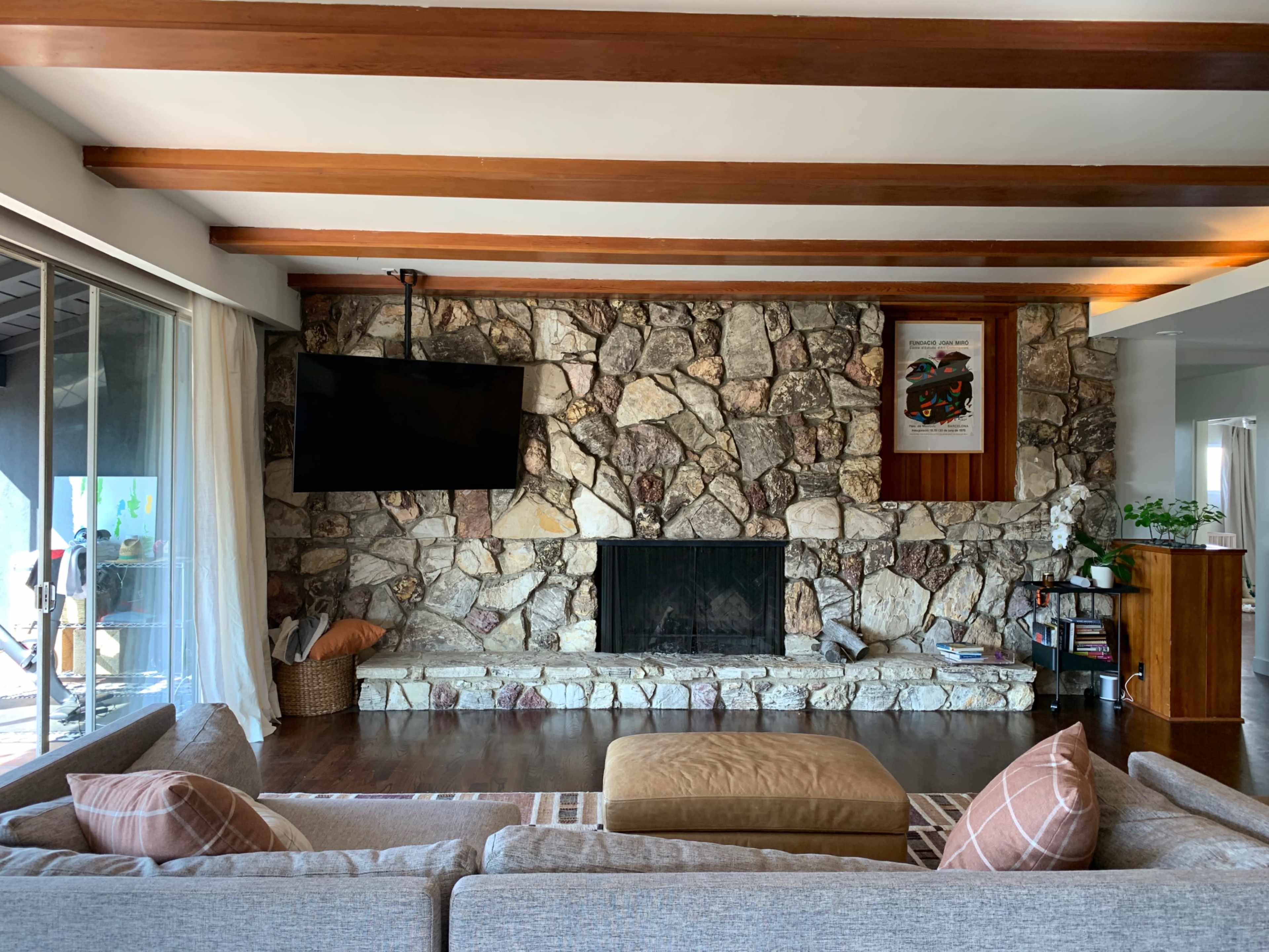 The image shows a living room featuring a stone fireplace, a wooden beam ceiling, a large television mounted on the wall, and a seating area with a flat sofa and an ottoman.