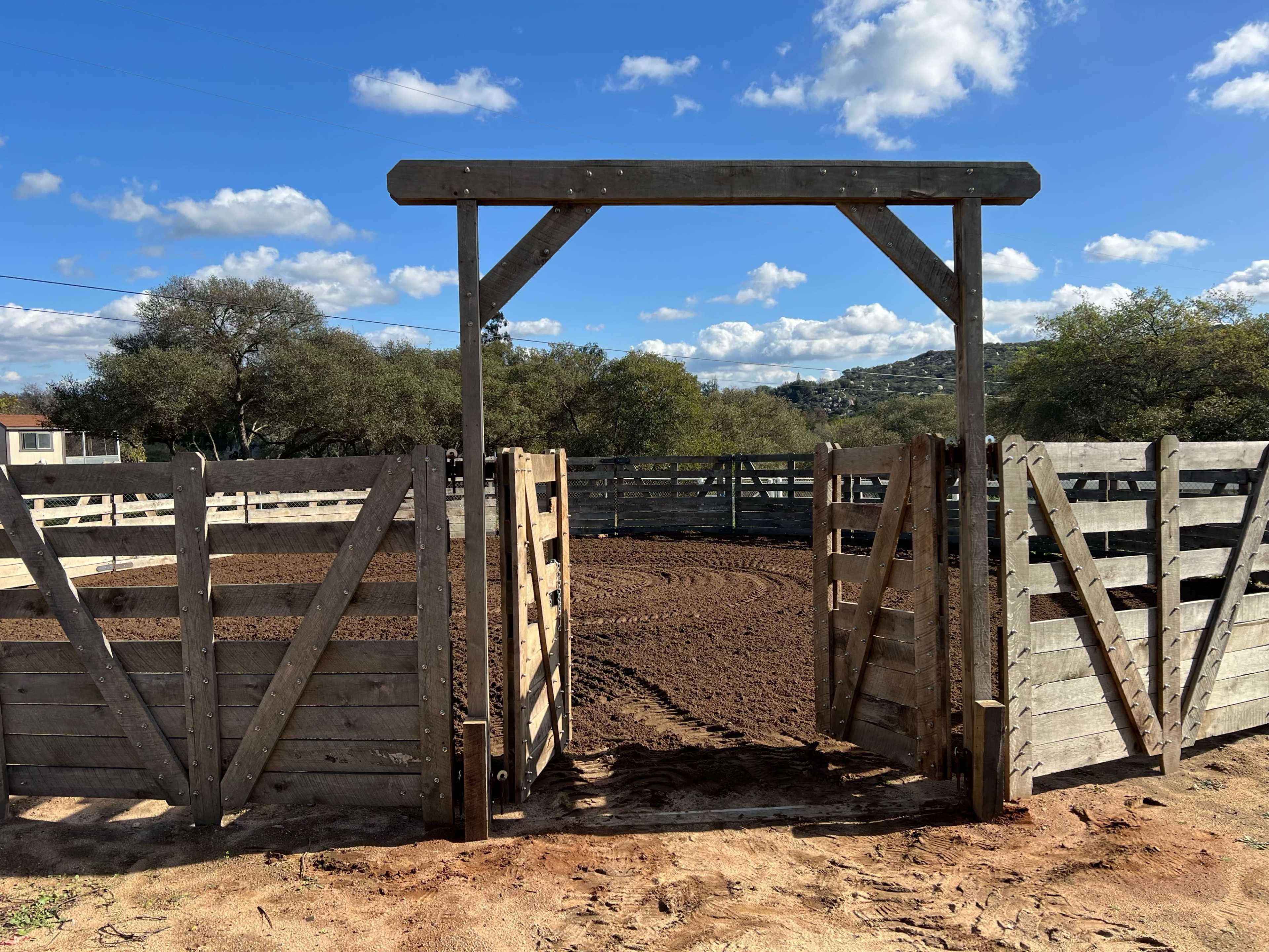 A wooden gate stands open at the entrance of a circular pen surrounded by wooden fencing under a partly cloudy sky.
