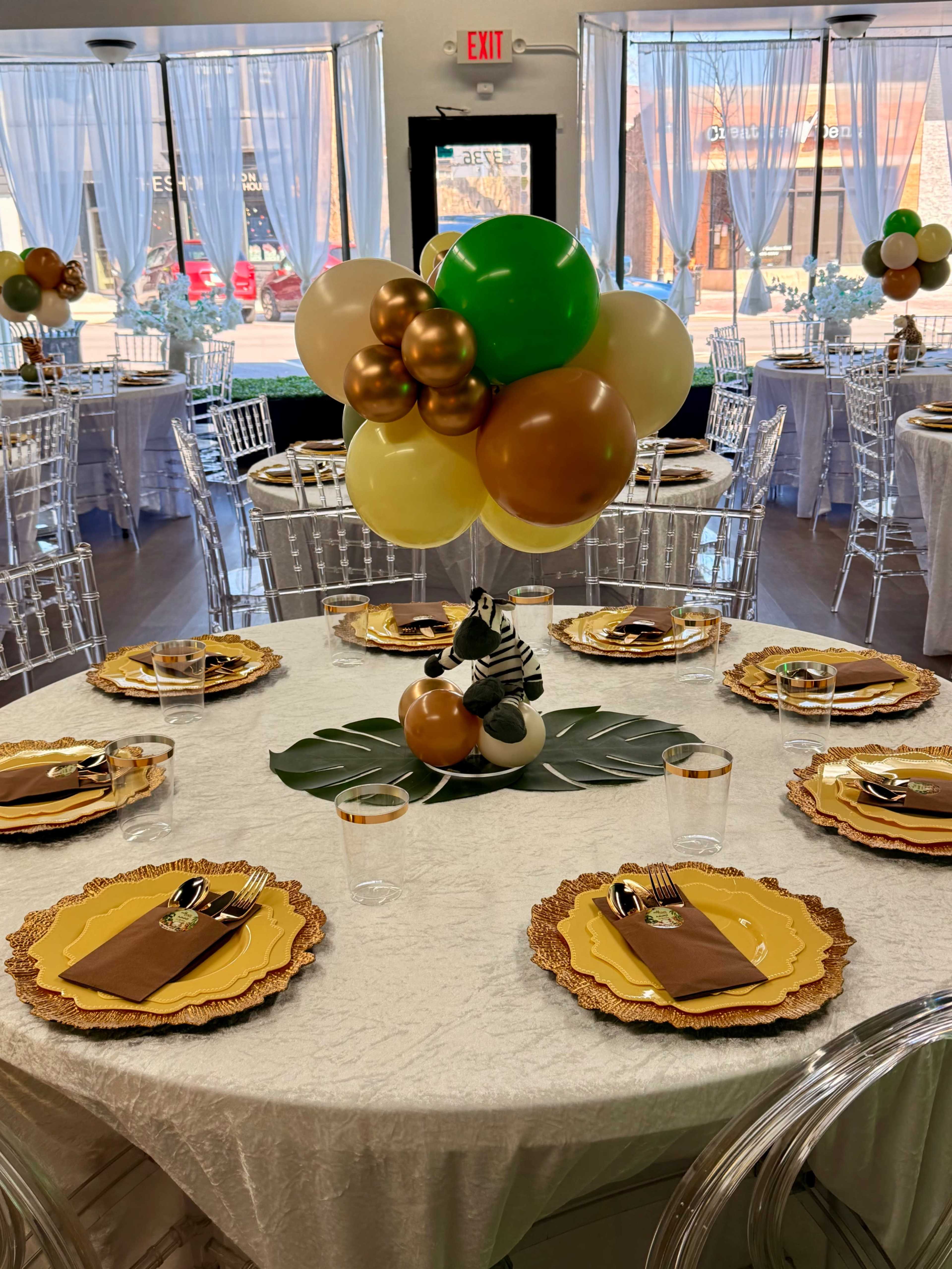 A decorated banquet room features round tables set with gold and brown tableware, surrounded by clear chairs and adorned with a colorful balloon centerpiece.