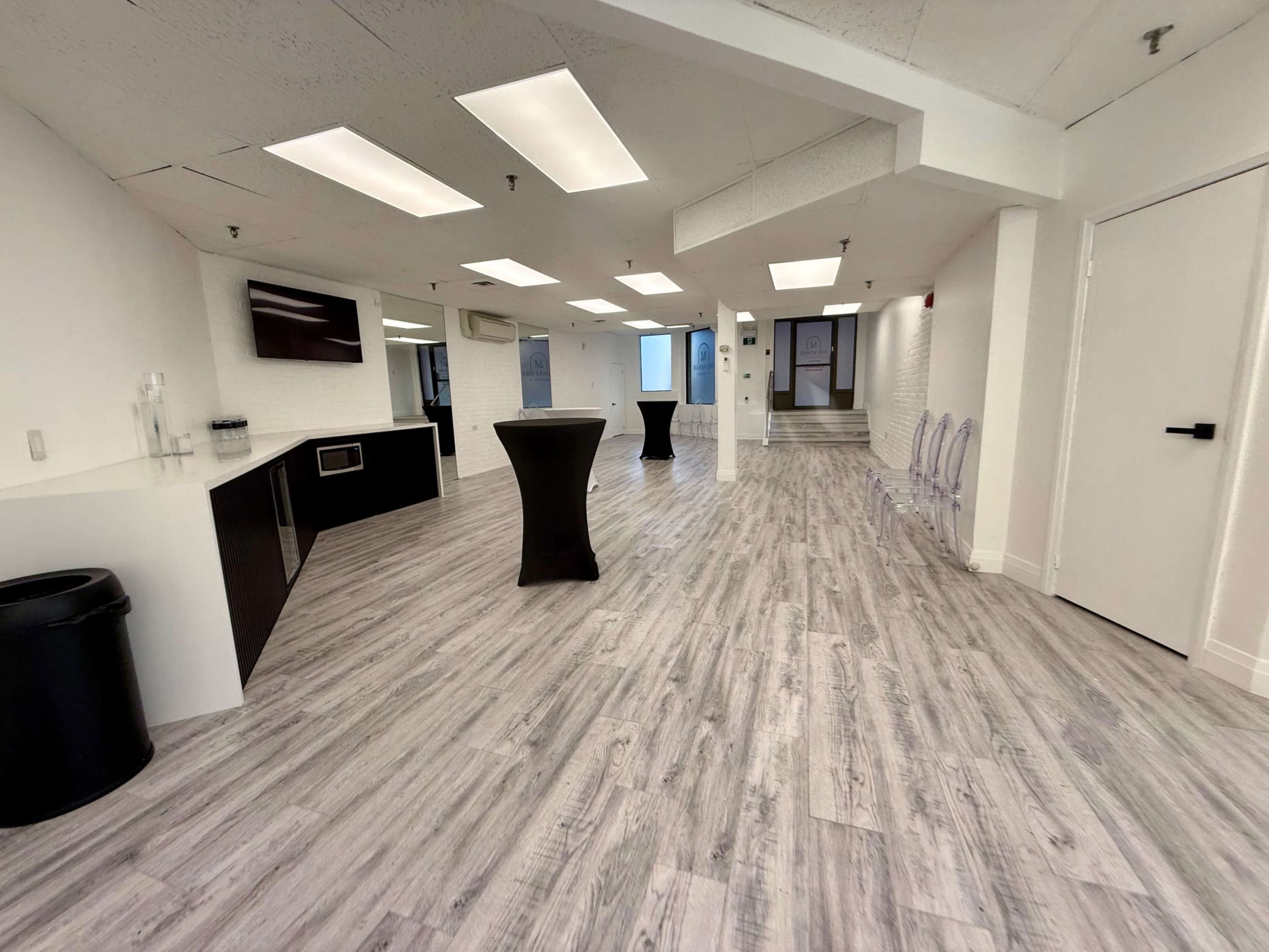 The image depicts a spacious, modern interior with light wood flooring, a kitchenette area, and clear plastic chairs scattered throughout the room.