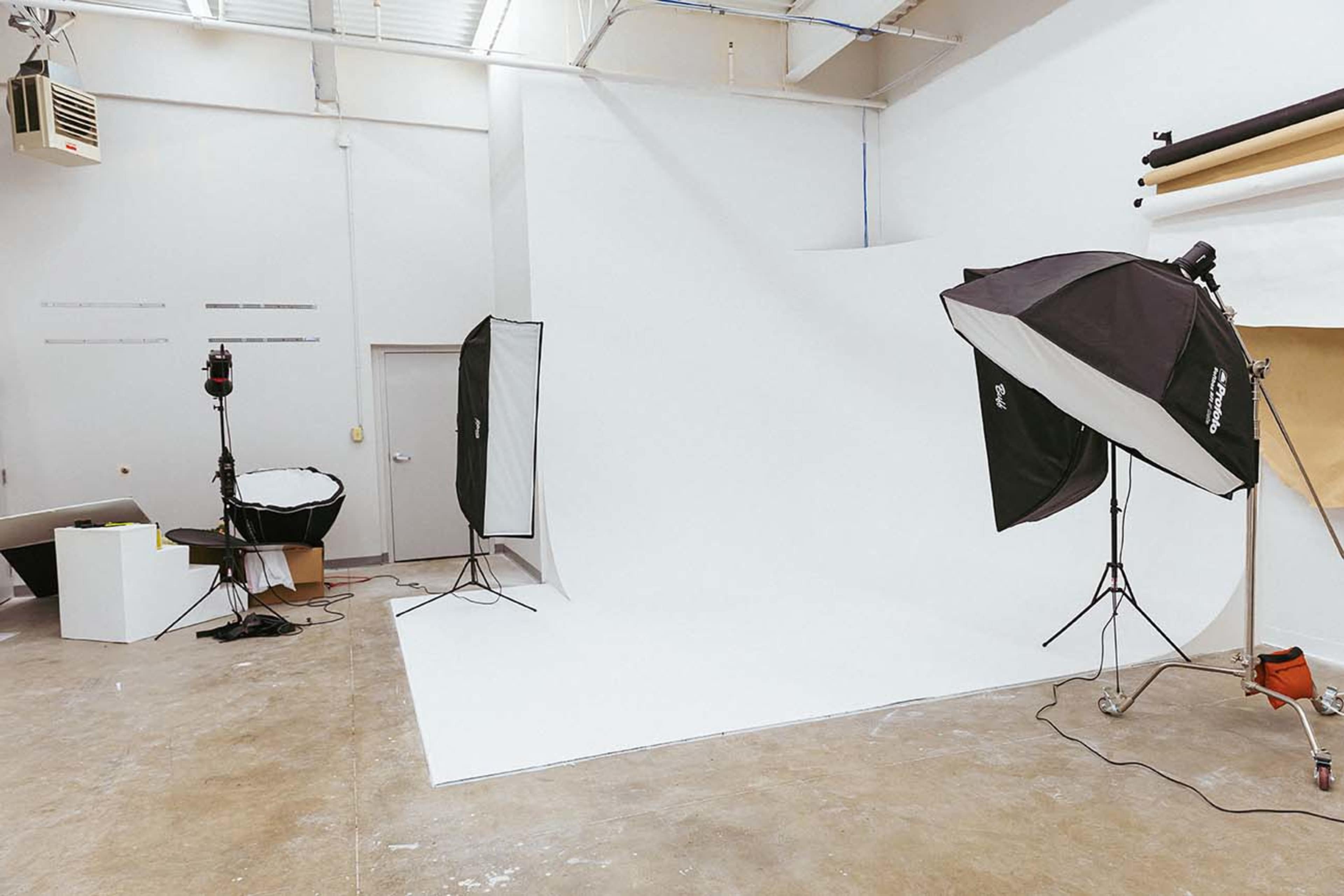 The image shows a photography studio with a large white backdrop, lighting equipment, and a few props arranged on the floor.