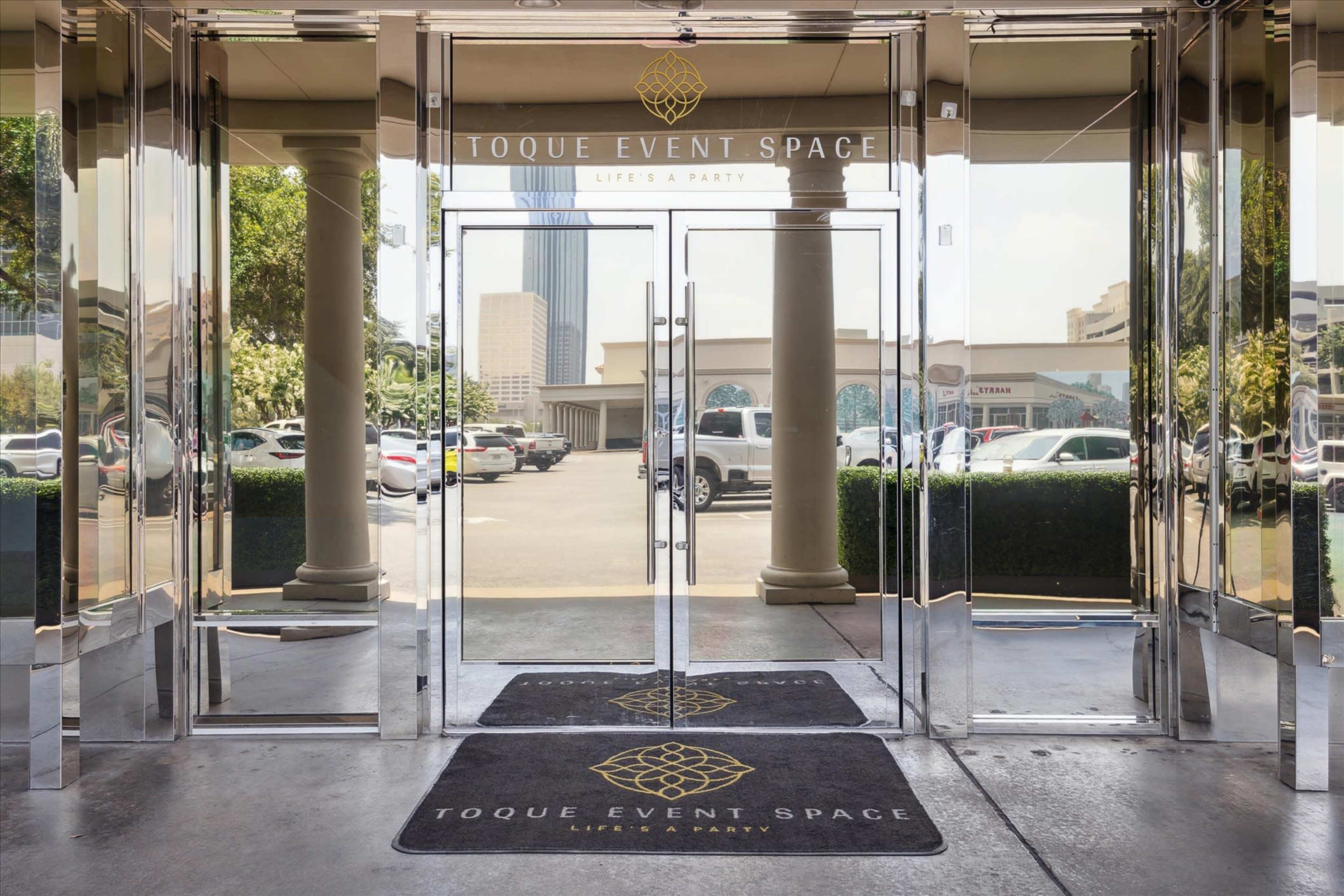 The entrance of Toque Event Space, featuring glass doors and a carpet with the venue's name, surrounded by parked cars and buildings in the background.