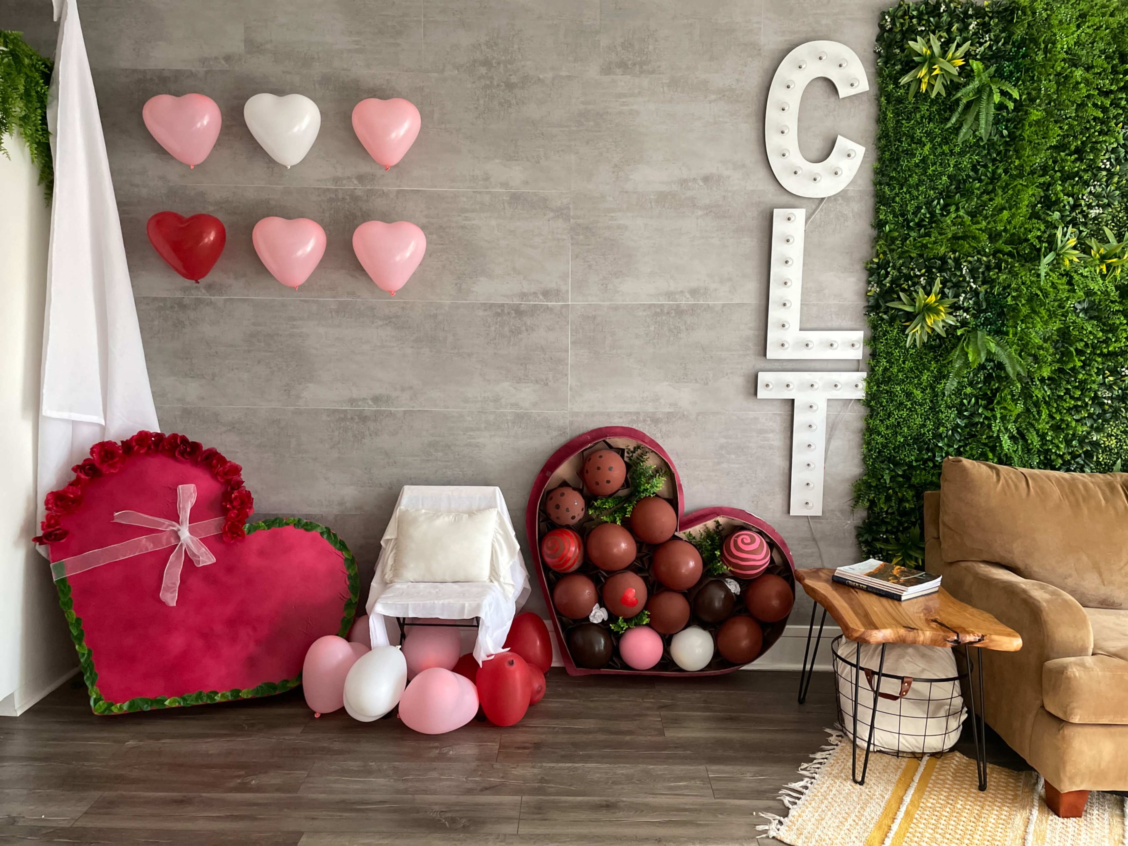 A decorated room with a gray wall featuring heart-shaped balloons, a large heart-shaped arrangement made of faux flowers and chocolates, and a cozy seating area with a brown sofa and a wooden side table.