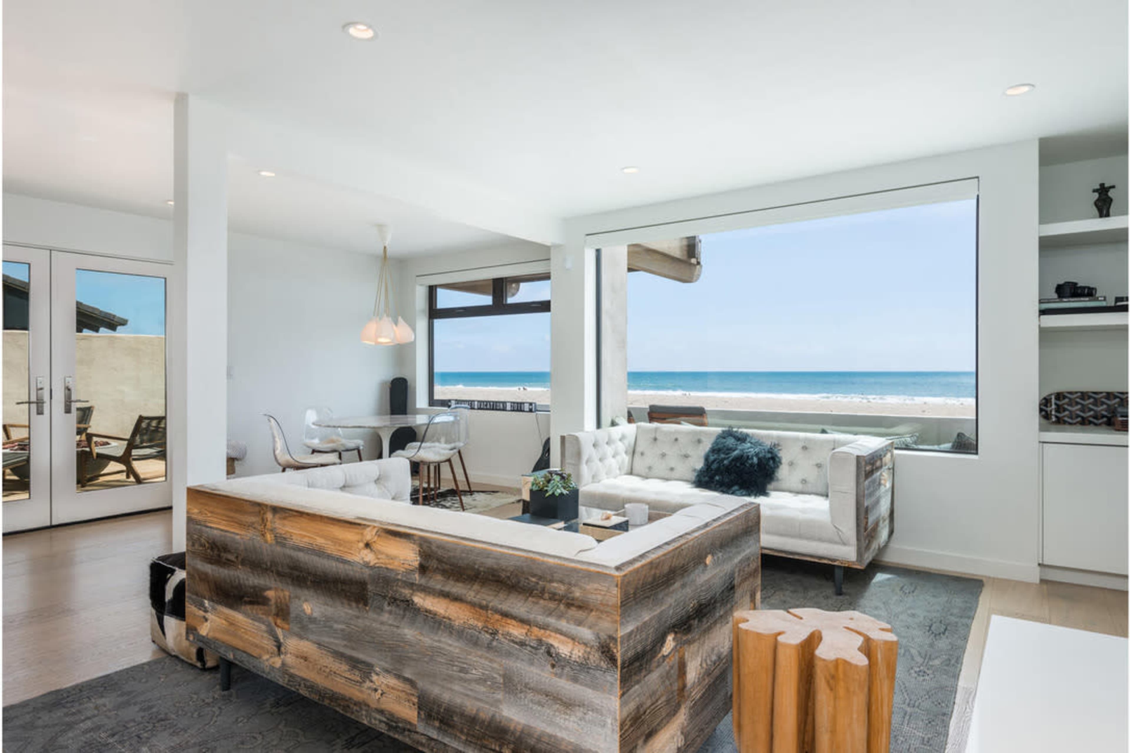 A modern living room features a wooden sofa, a small dining area, and large windows overlooking the ocean.