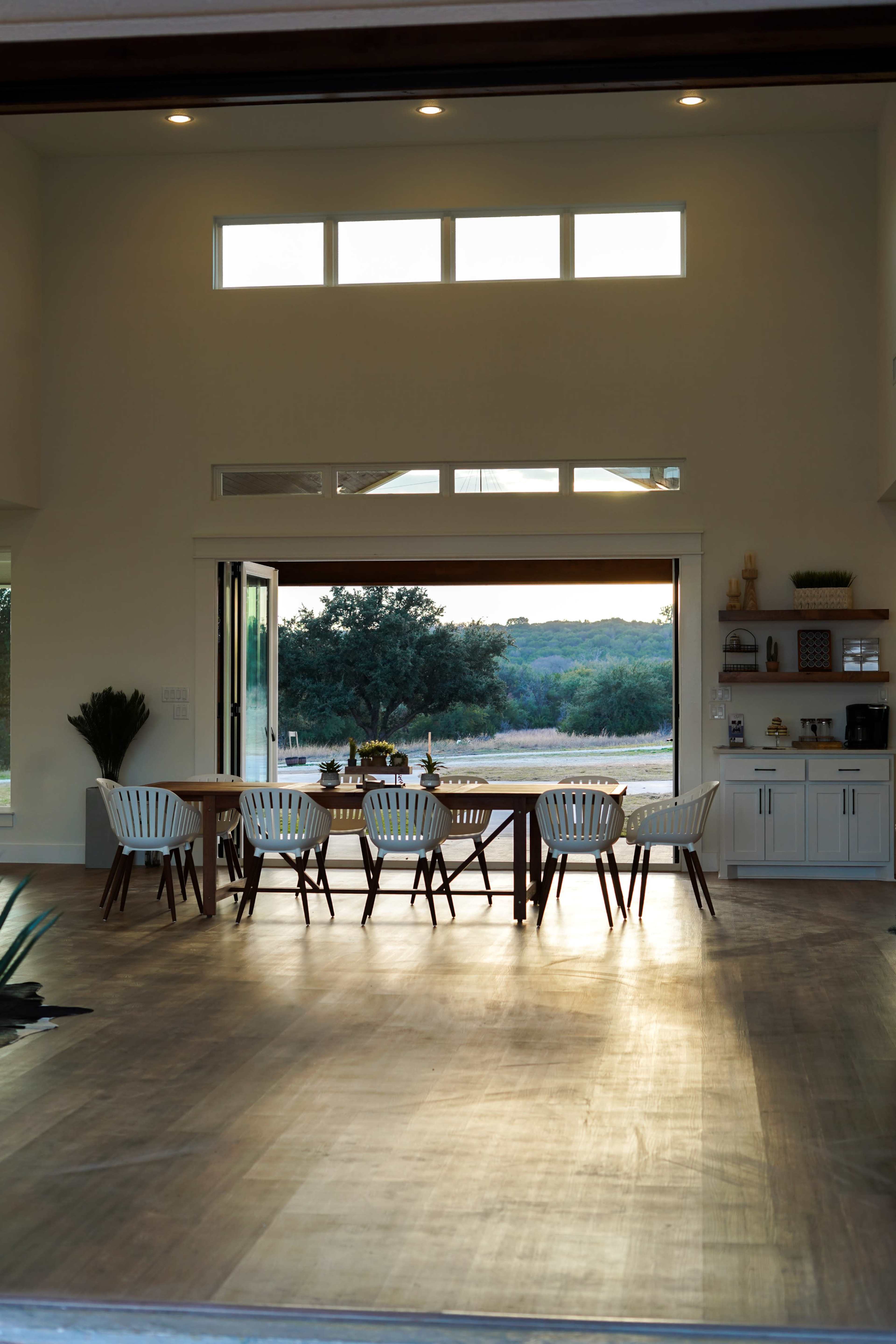 A dining area features a long wooden table surrounded by white chairs, with large windows offering a view of the outdoors.
