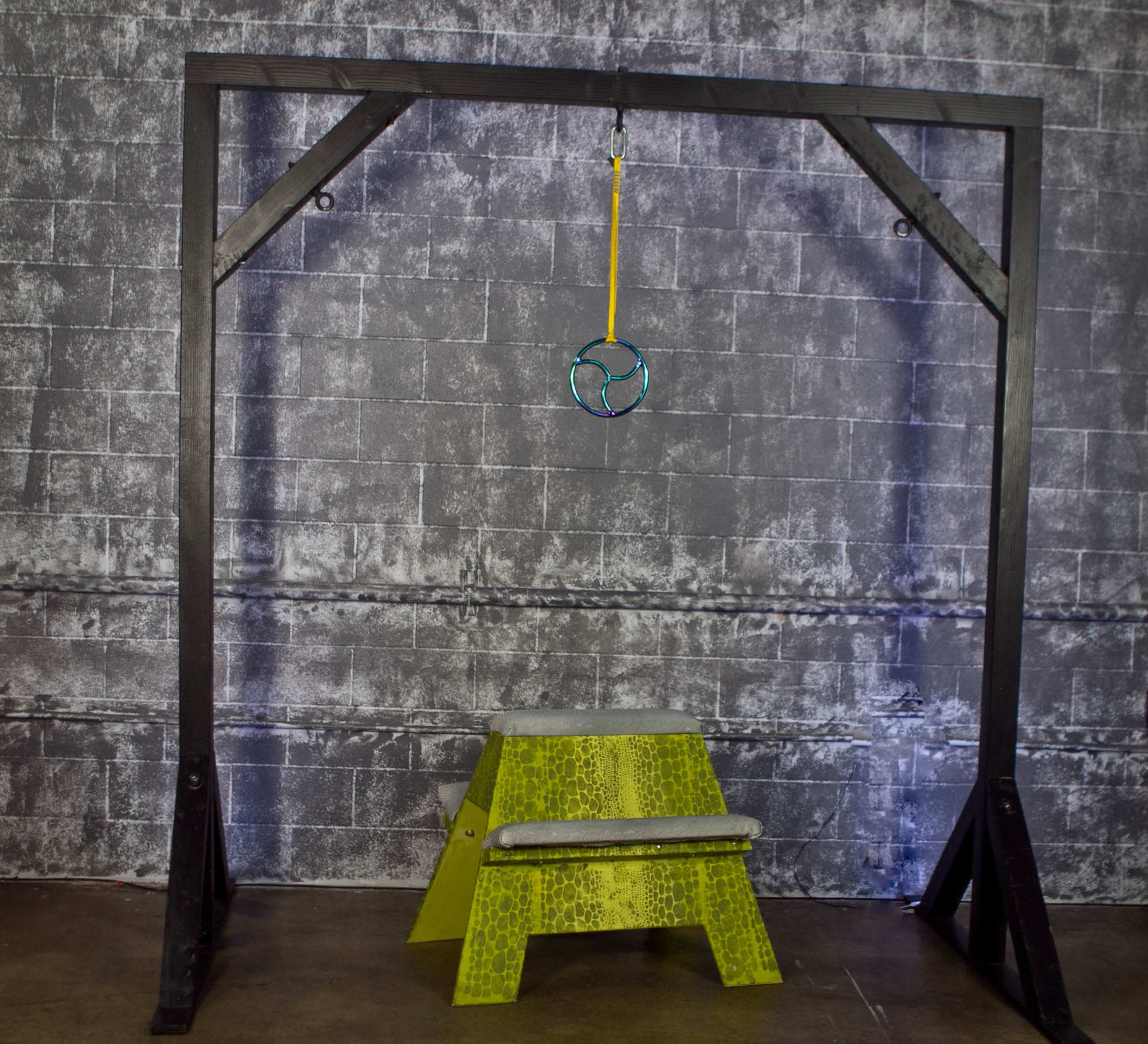 A black exercise apparatus with a hanging hoop and a yellow stool beneath it, set against a textured gray wall.