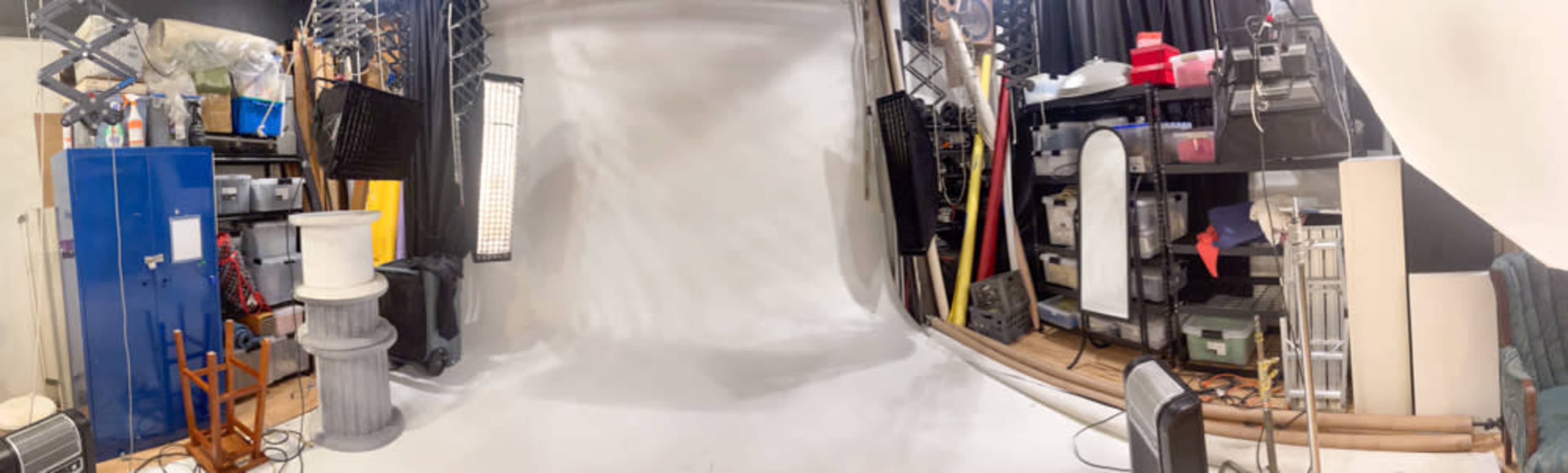 The image shows a photography studio with a white backdrop and various equipment stored along the walls.