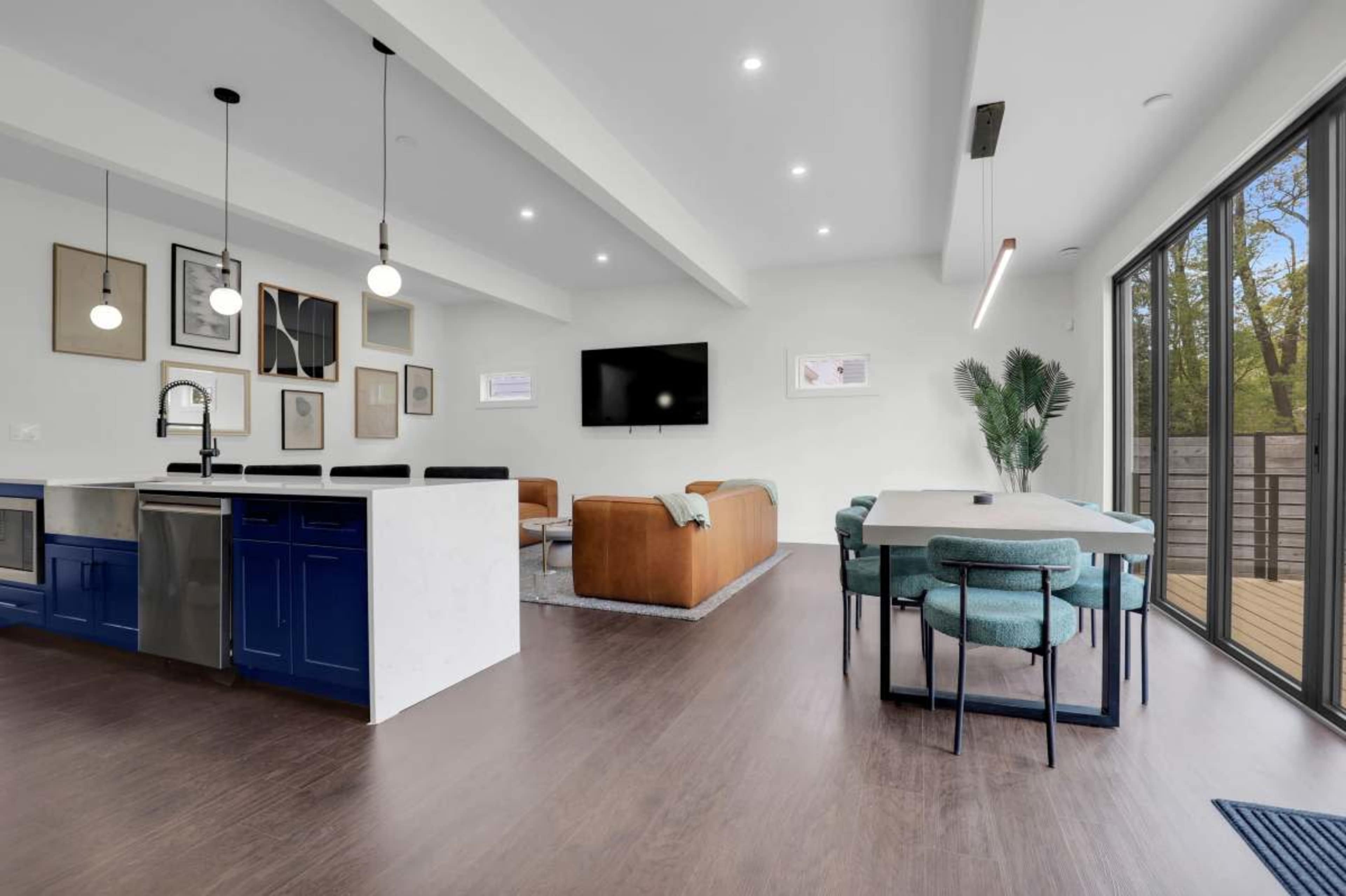 The image shows a modern, open-concept living space with a kitchen, dining area, and living room, featuring a mix of blue and brown furnishings and large windows leading to an outdoor deck.