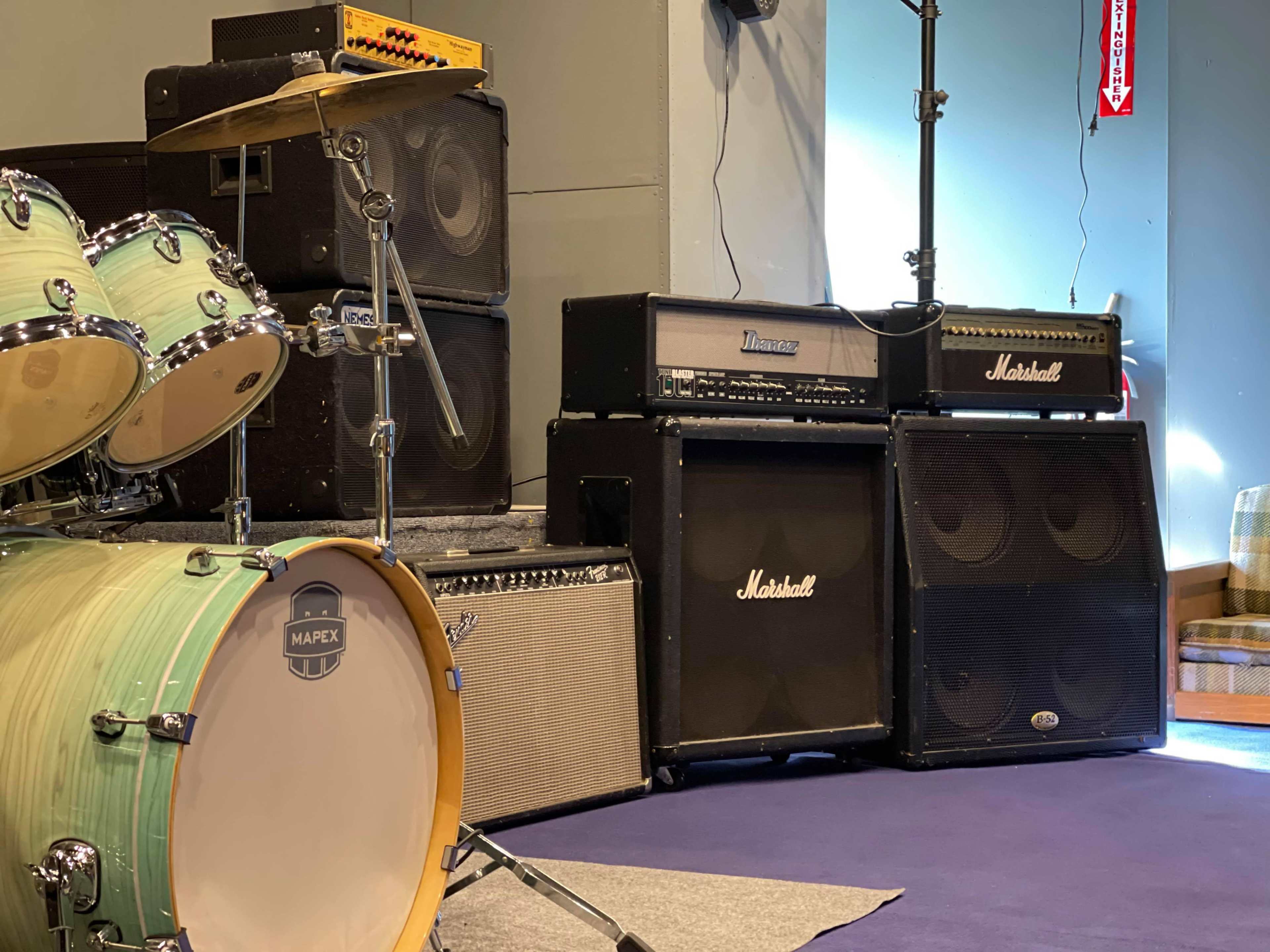 A collection of amplifiers stacked next to a drum set in a rehearsal space.