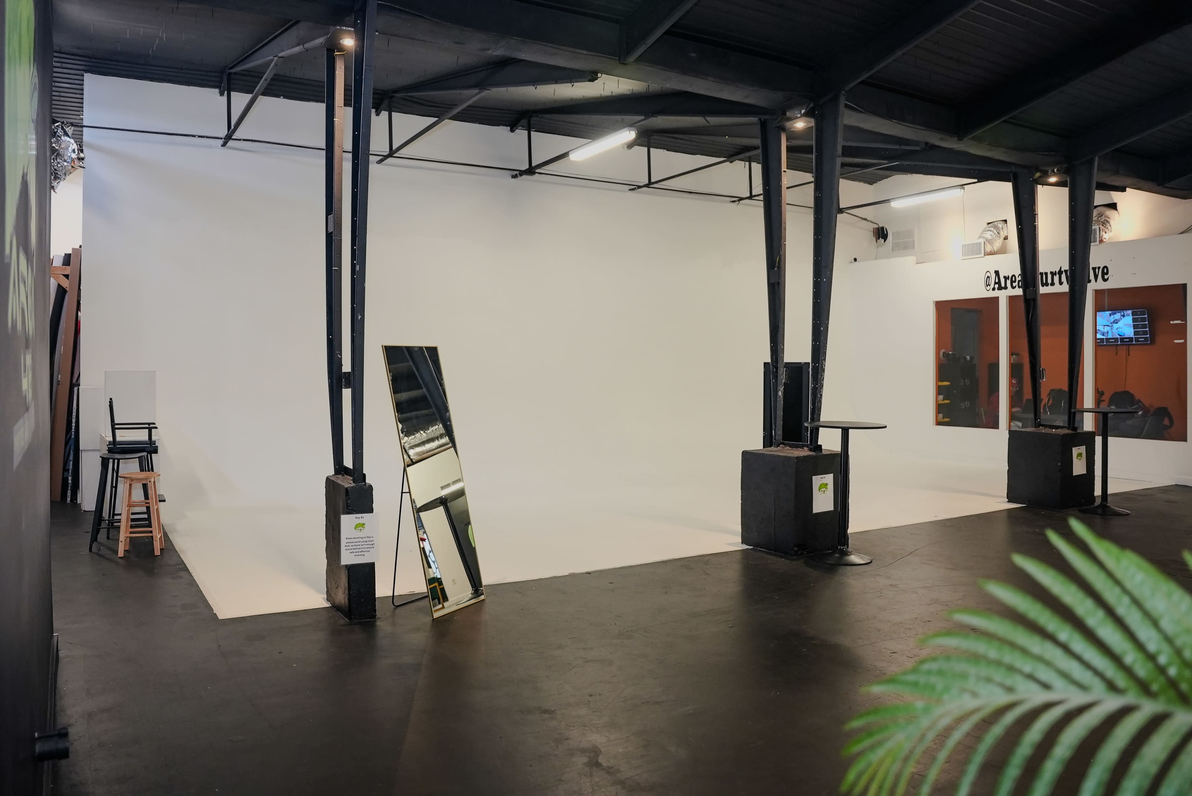 The image shows a spacious, minimalist studio with a large white backdrop and black flooring, featuring a mirror and some furniture in the corner.
