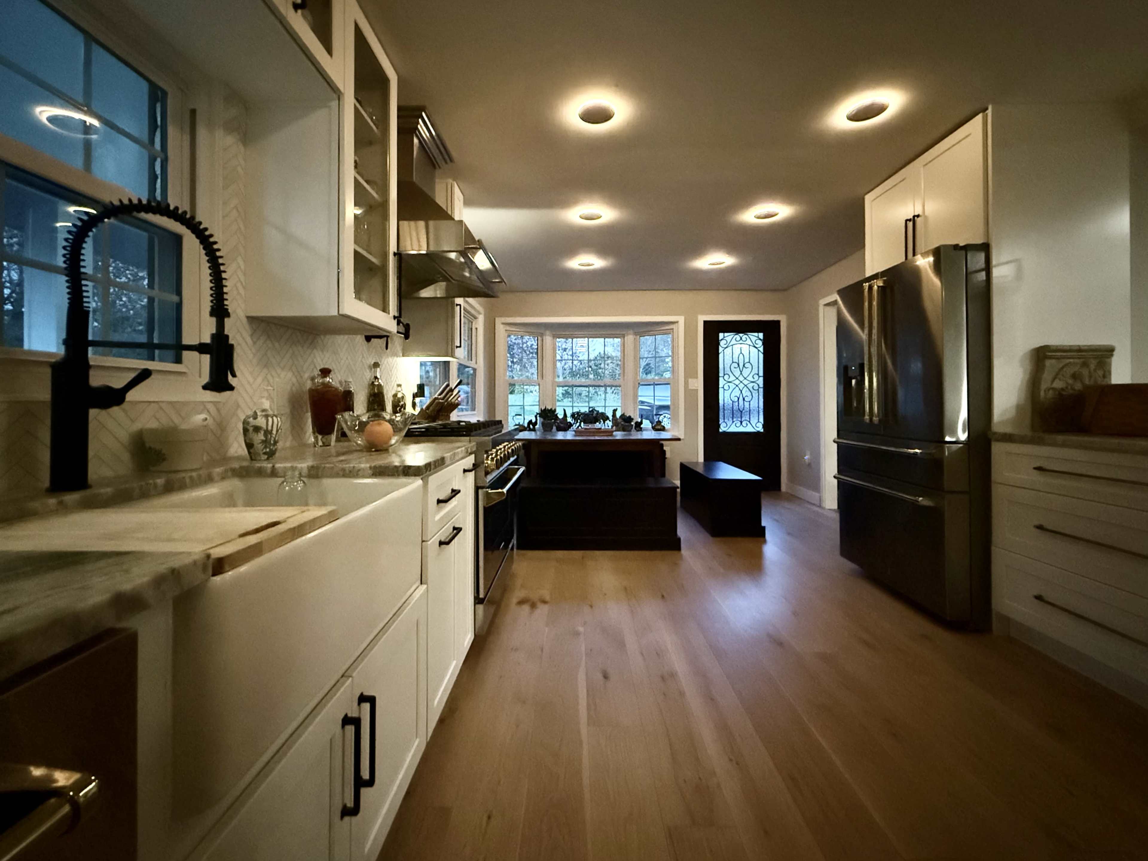 The image shows a modern kitchen with white cabinets, a marble sink, stainless steel appliances, and large windows providing natural light.