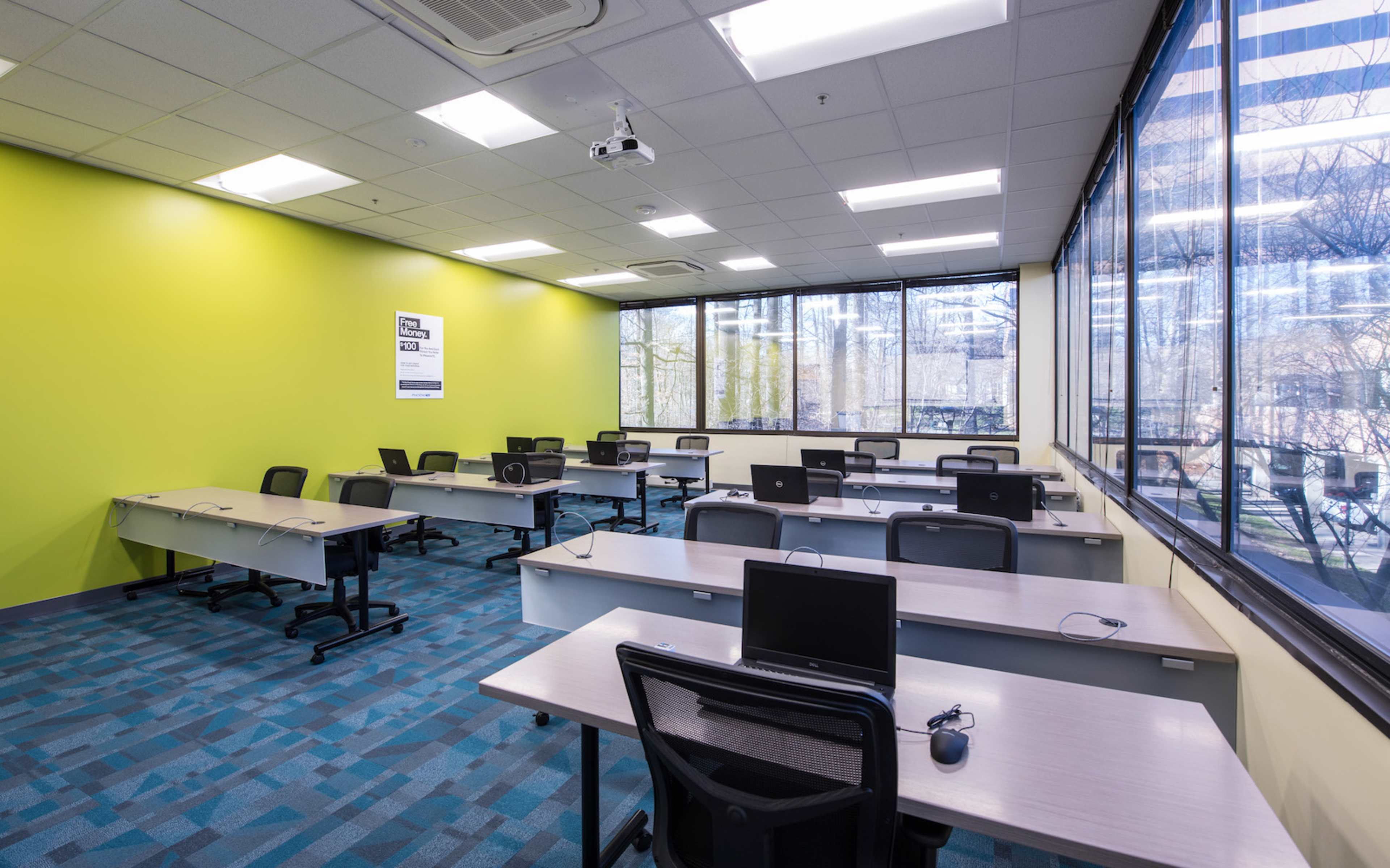 The image shows a bright classroom with multiple desks equipped with computers, large windows allowing natural light, and a green accent wall.