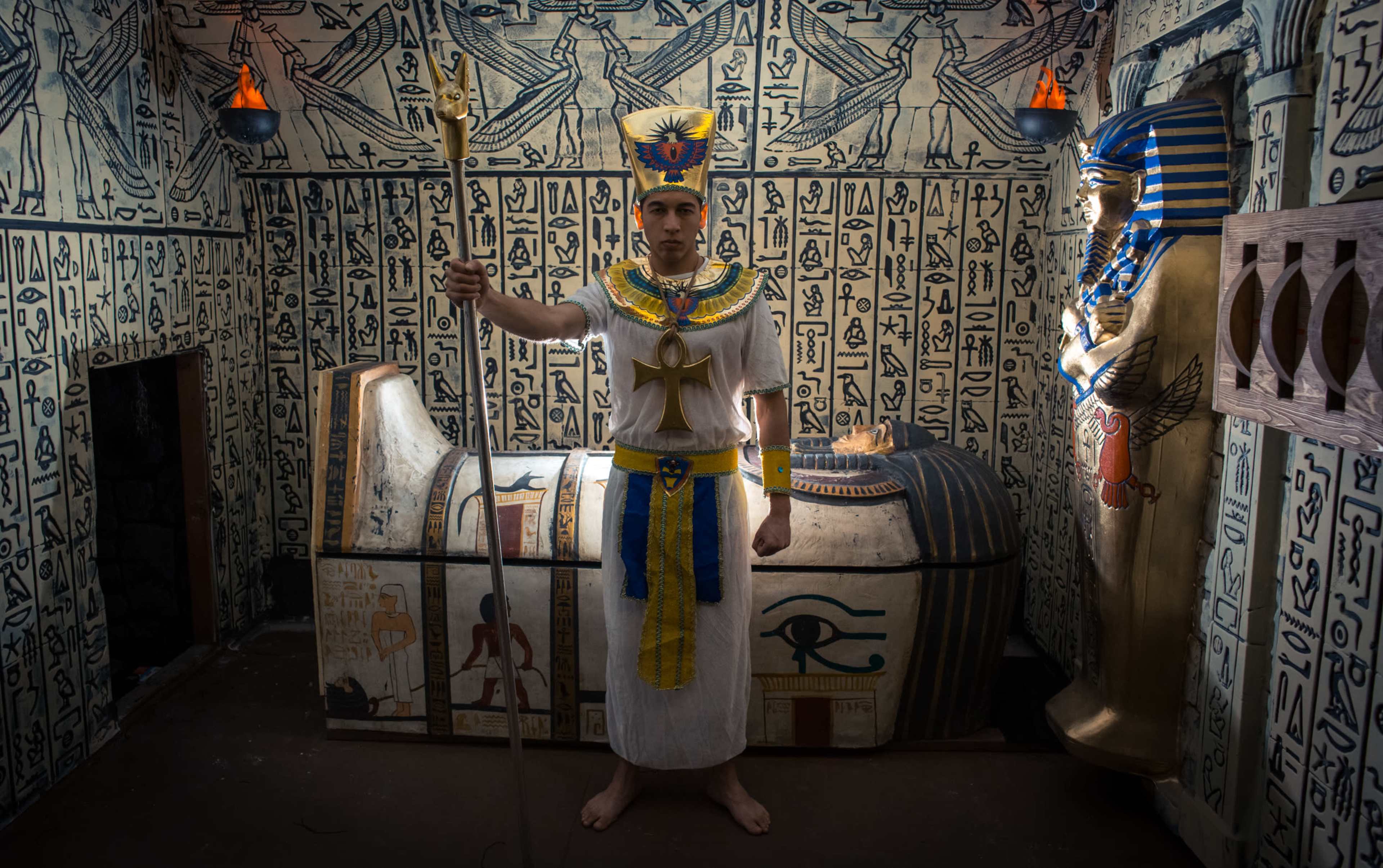 A person dressed in ancient Egyptian attire stands in a room decorated with hieroglyphics, next to a sarcophagus and a statue of a pharaoh.