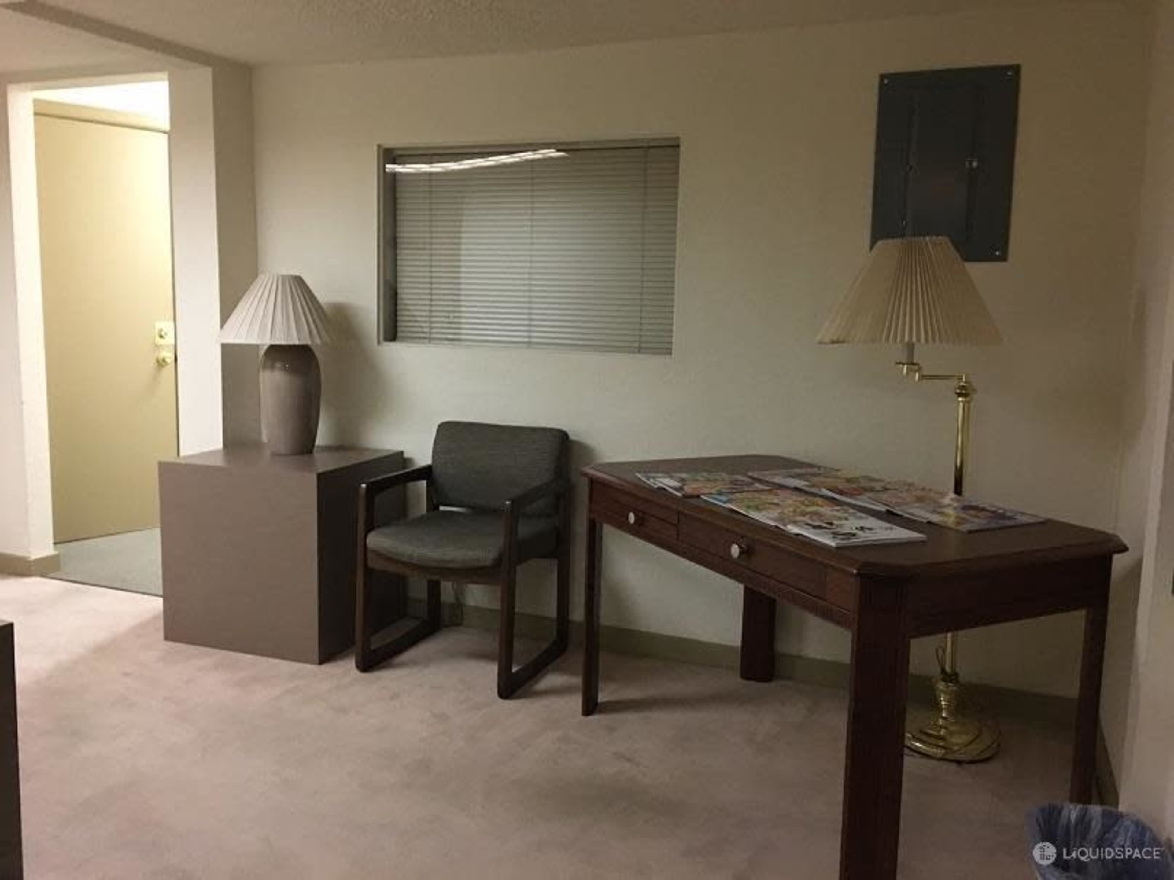A plain room with a chair and a wooden desk, accompanied by a lamp and a small side table.