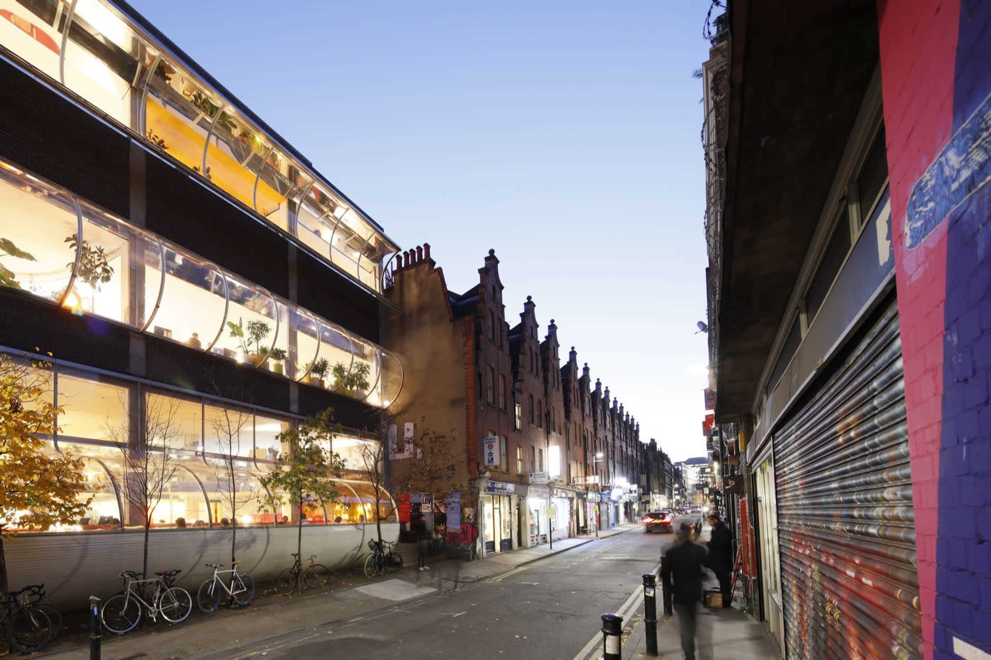 The image shows a street scene with a modern glass building on one side and historic brick buildings on the opposite side, with bicycles parked along the curb and soft evening light illuminating the area.