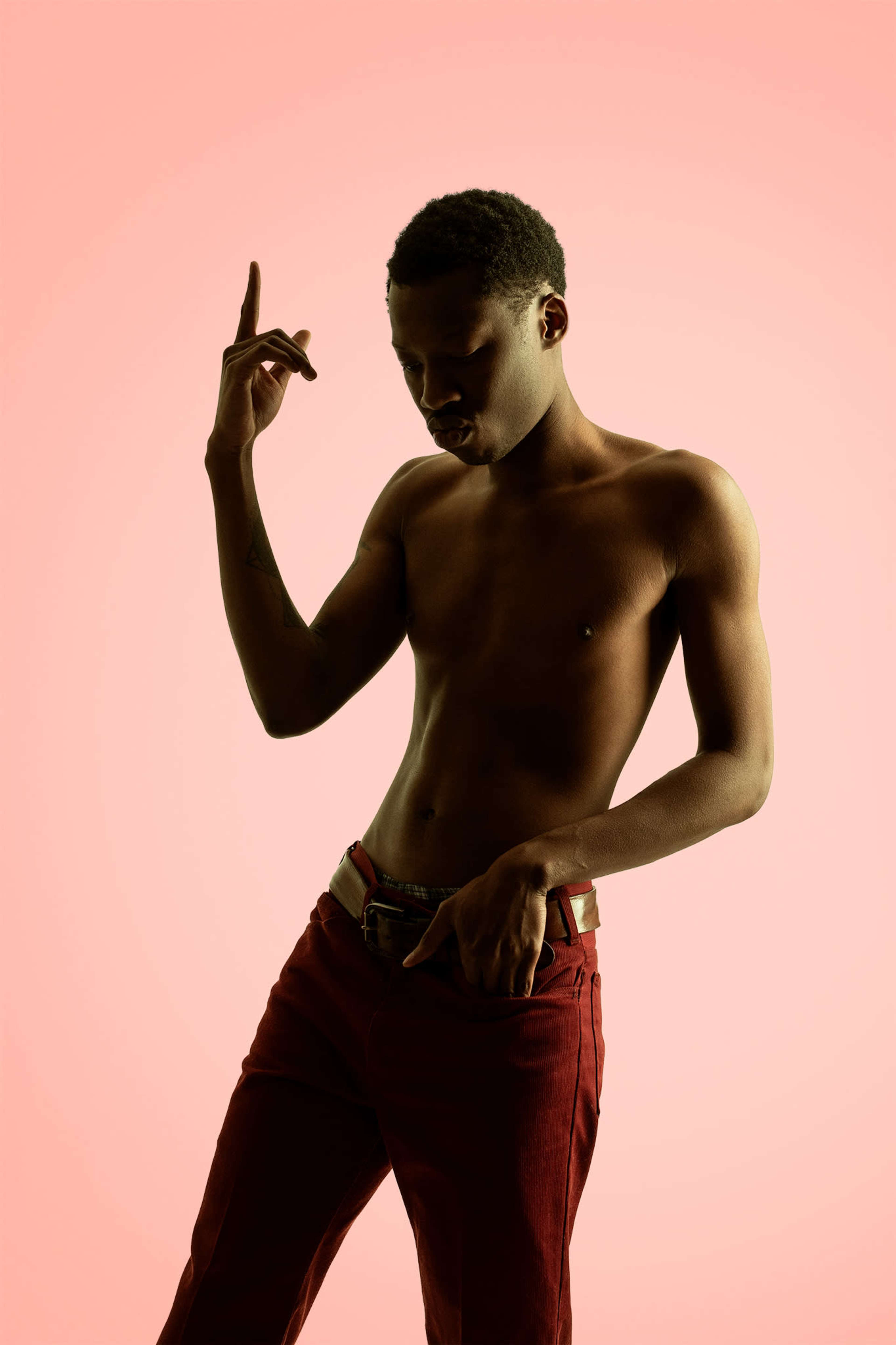 A person poses with one hand raised and the other holding their waistband, set against a pink background.