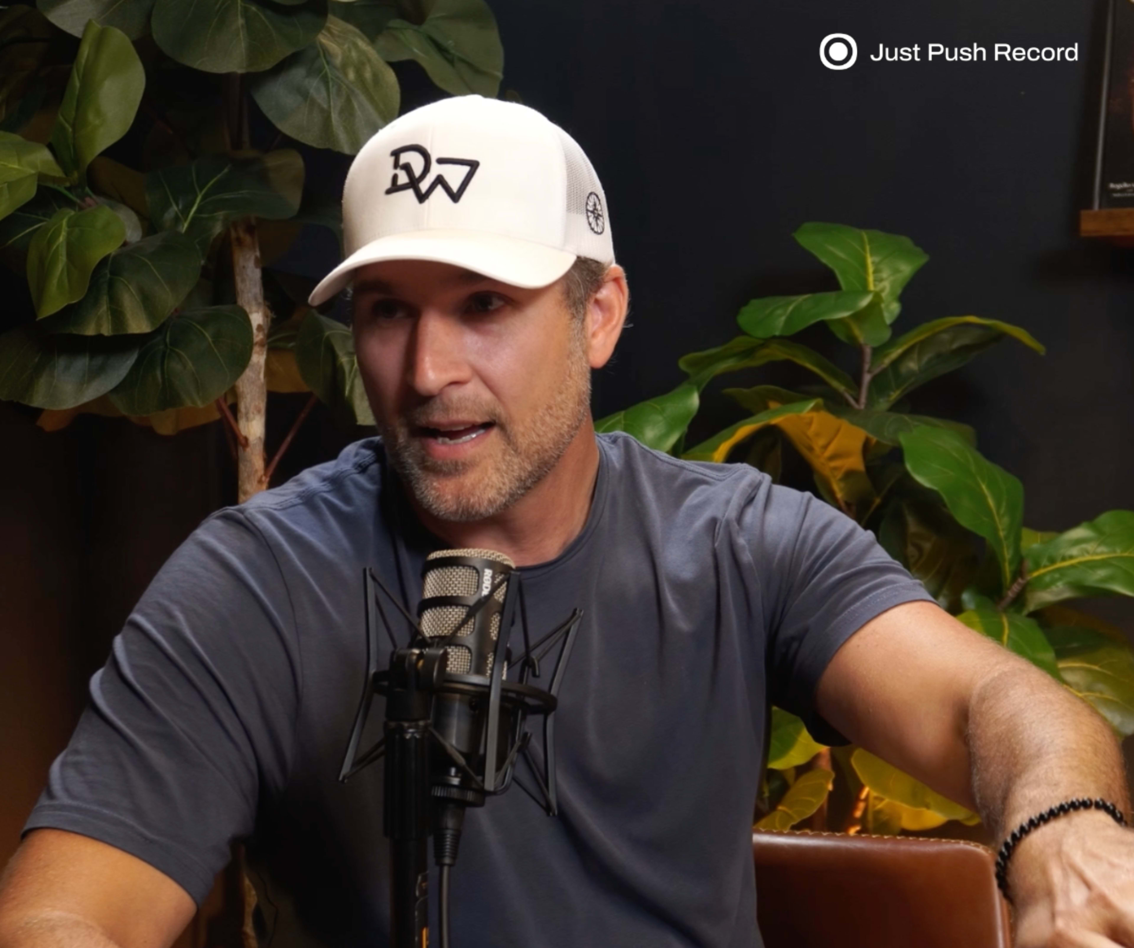 A man wearing a white baseball cap and a gray T-shirt speaks into a microphone while seated in front of greenery.