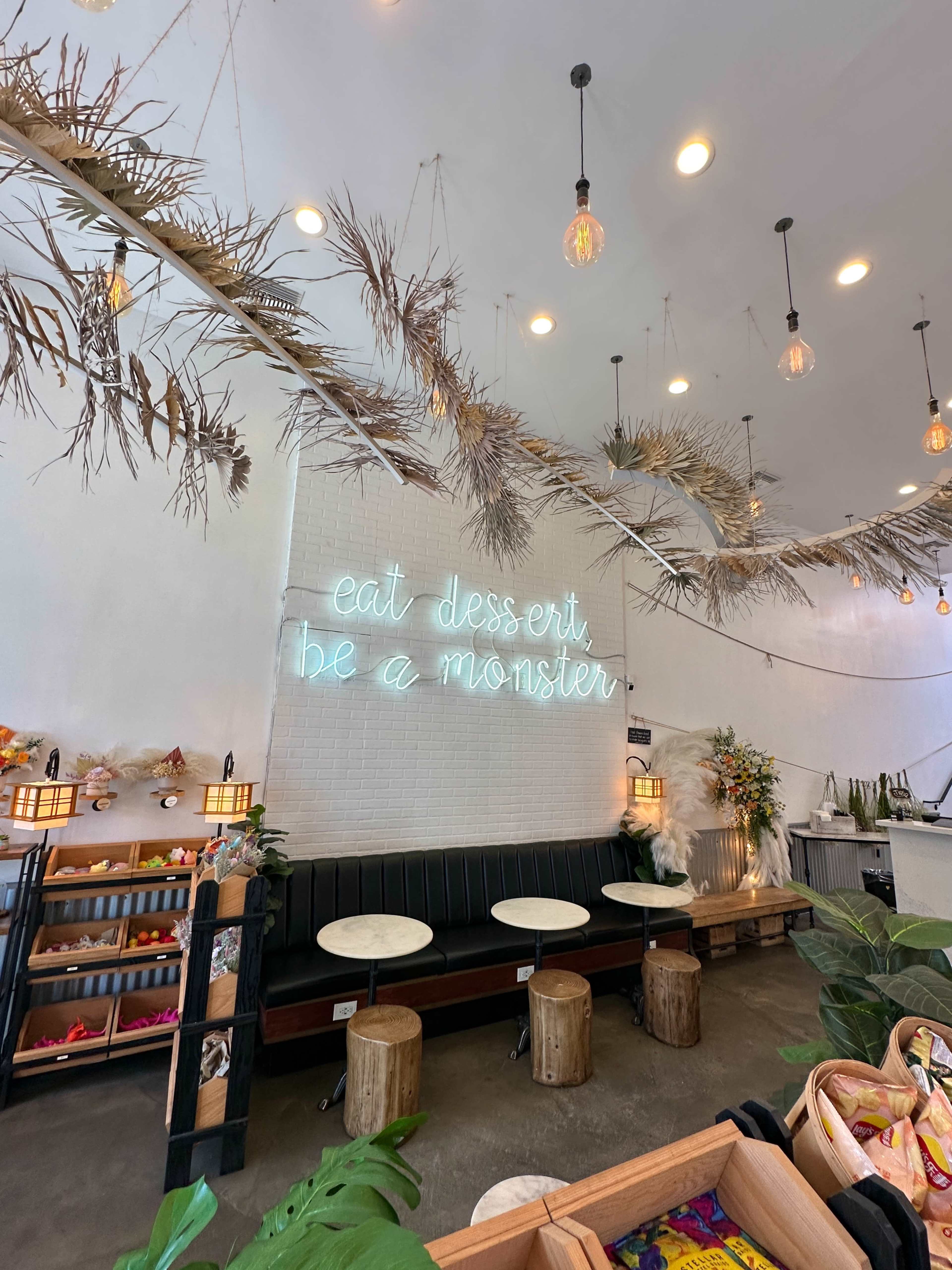 The interior of a café features a neon sign that reads "eat dessert be a monster," with hanging decorations and a seating area equipped with wooden stools and tables.