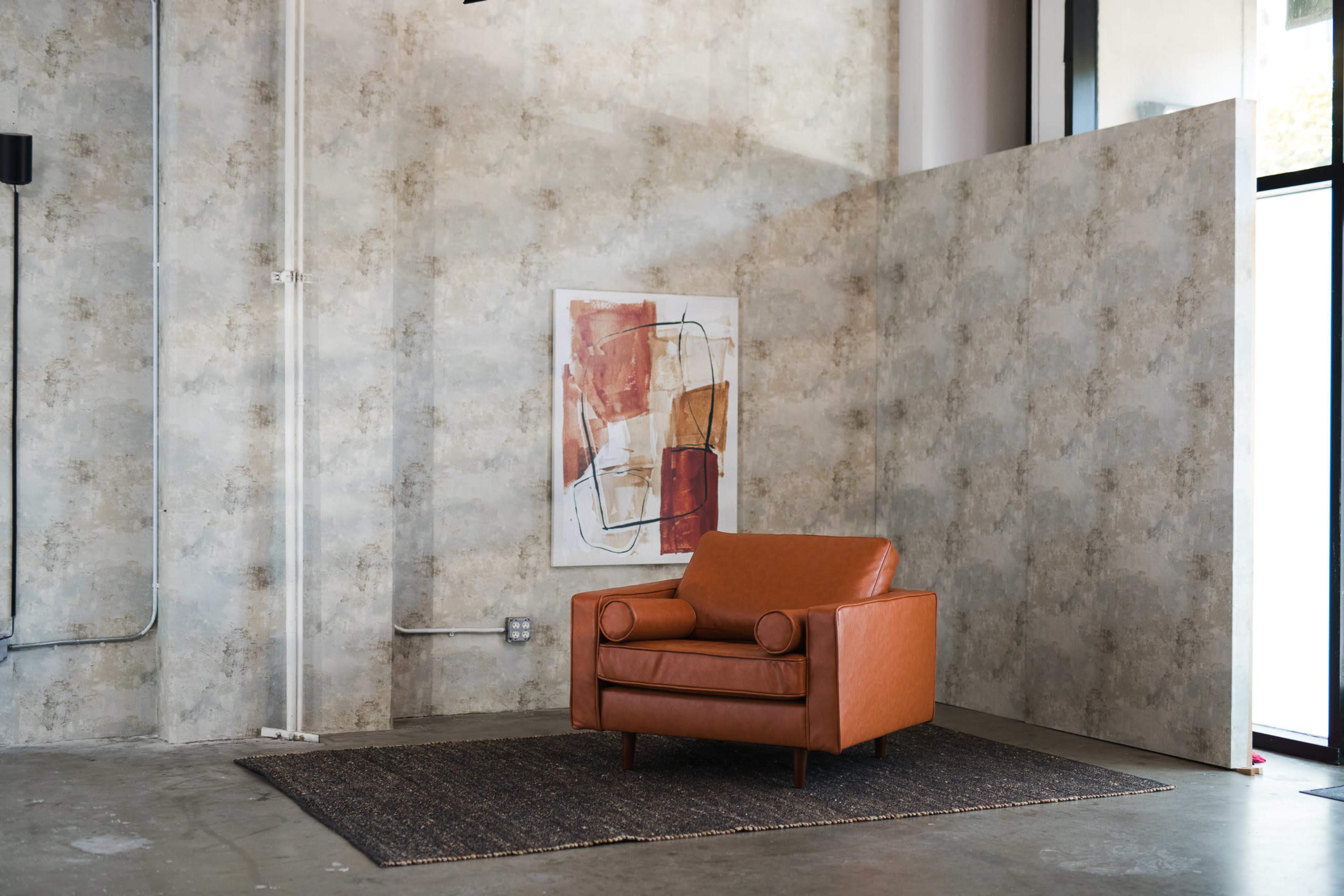 A brown leather armchair is positioned against a textured wall with an abstract painting above it, on a woven rug in a spacious room with large windows.