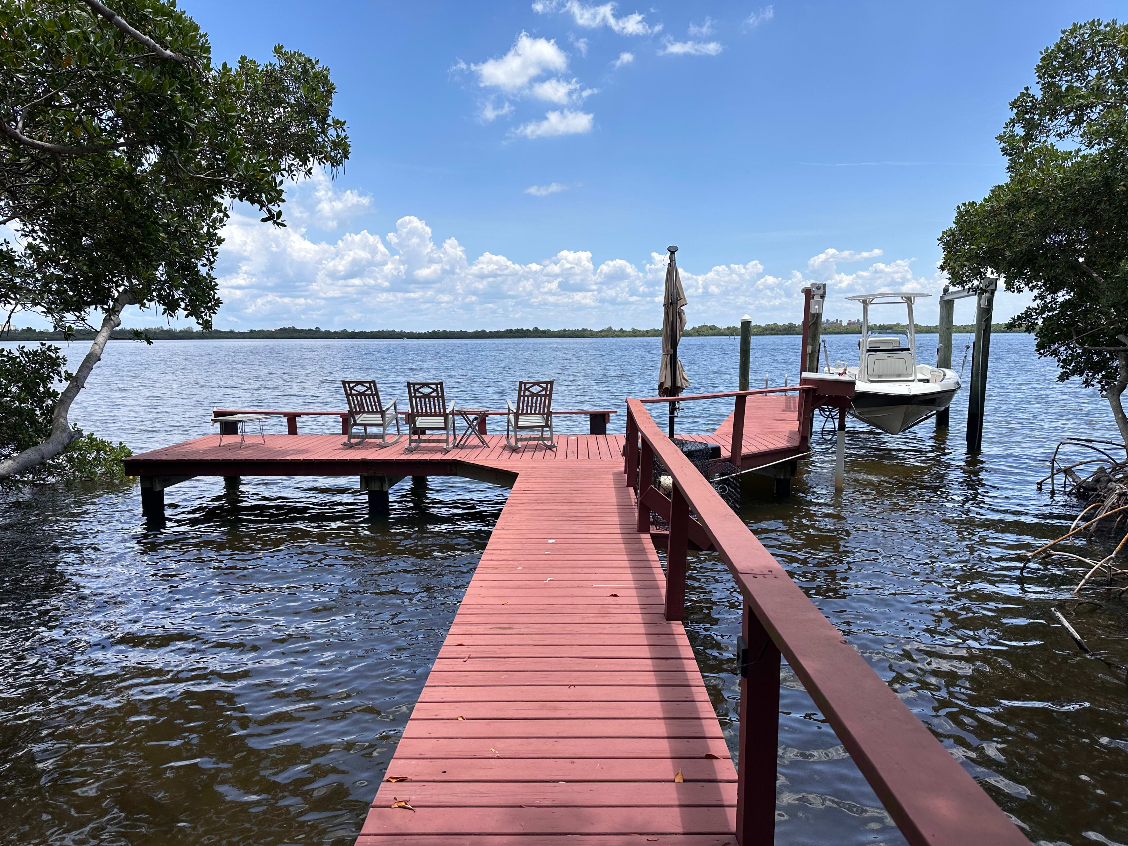 A red wooden dock extends into a body of water, with chairs set up at the end and a small boat moored beside it.