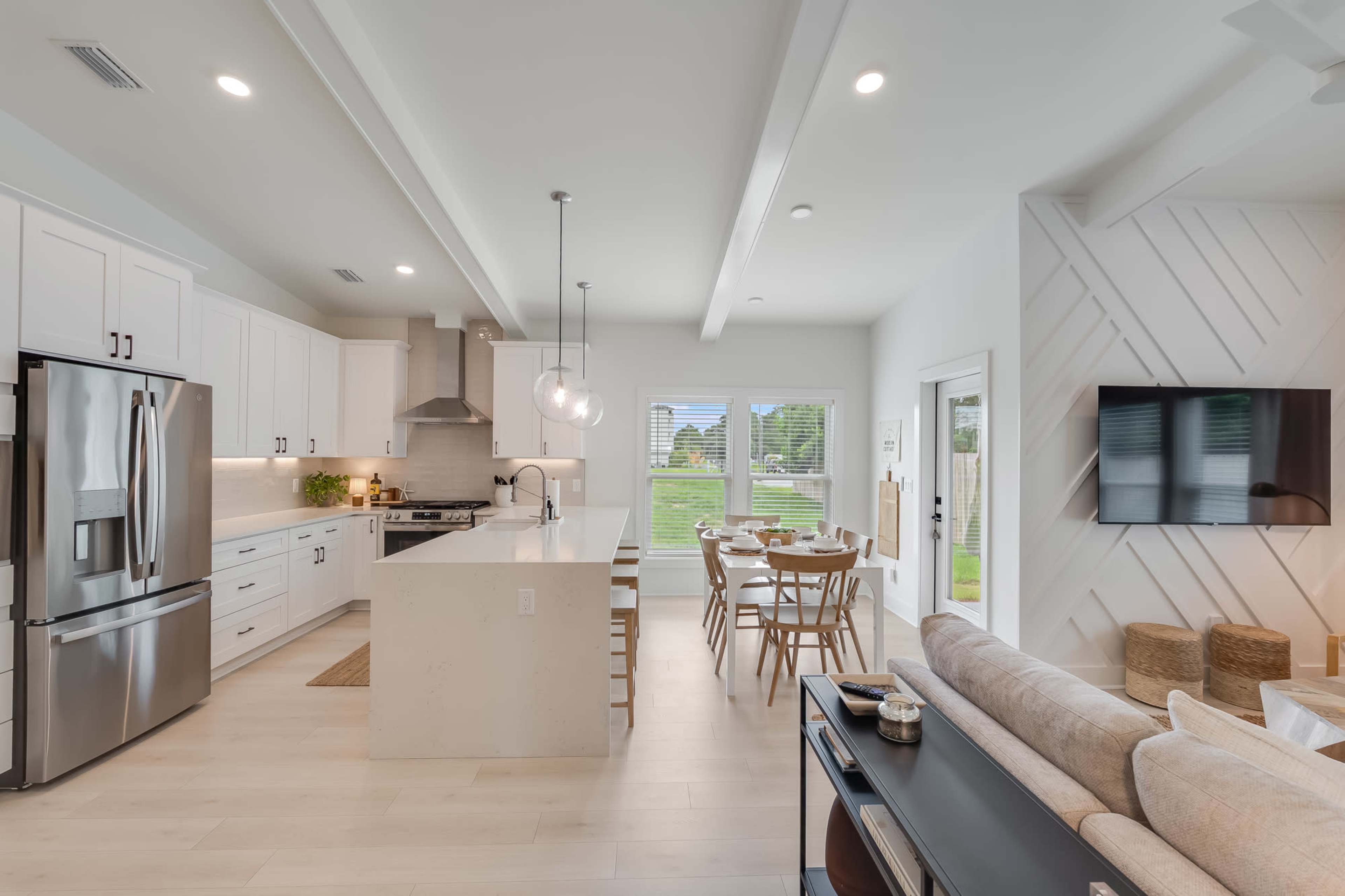 The image shows a modern kitchen and living area with white cabinetry, stainless steel appliances, and a dining table near large windows.