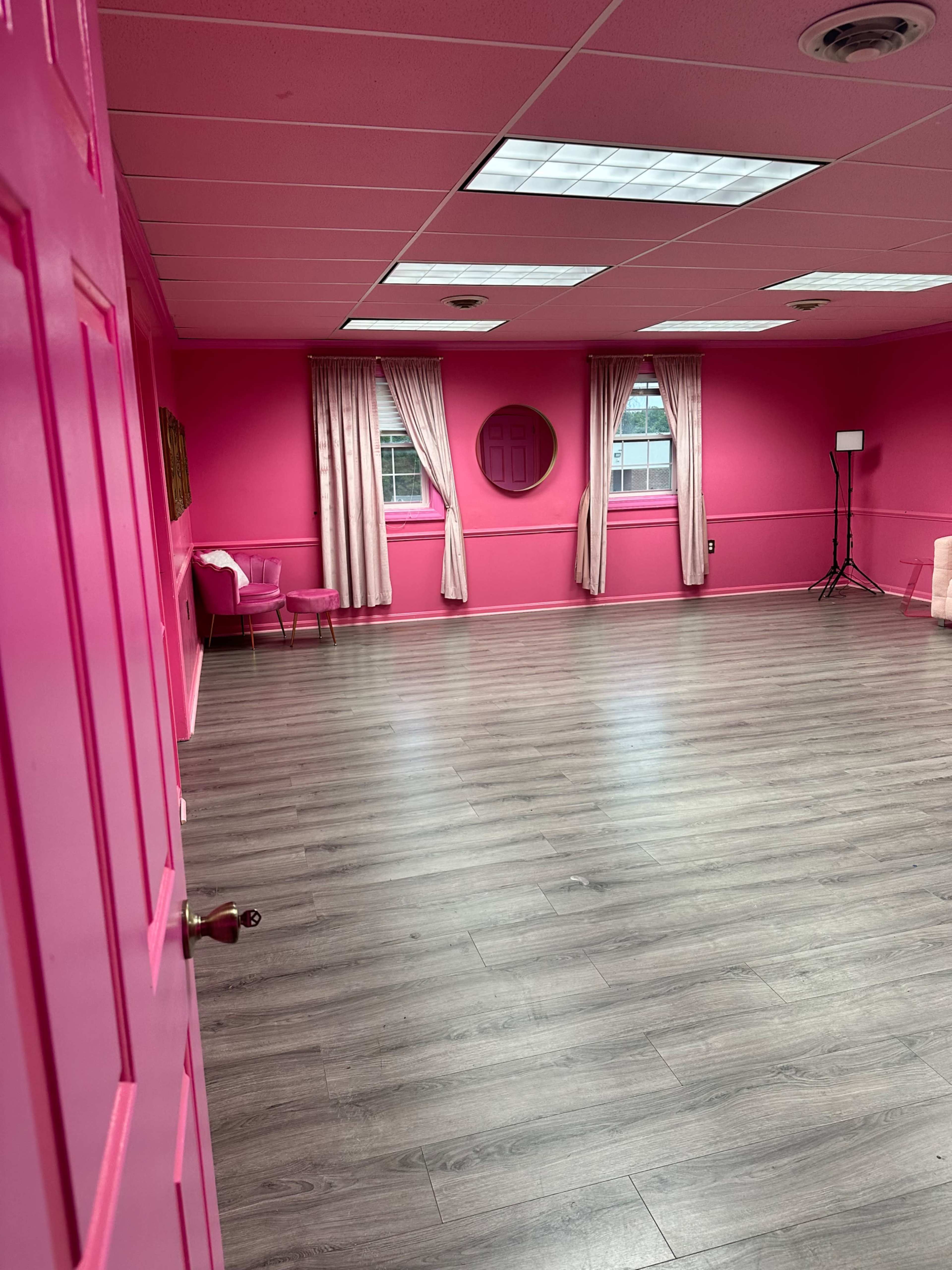 The image shows a brightly painted pink room with wooden flooring, featuring curtains, a round mirror on the wall, and a small seating area.