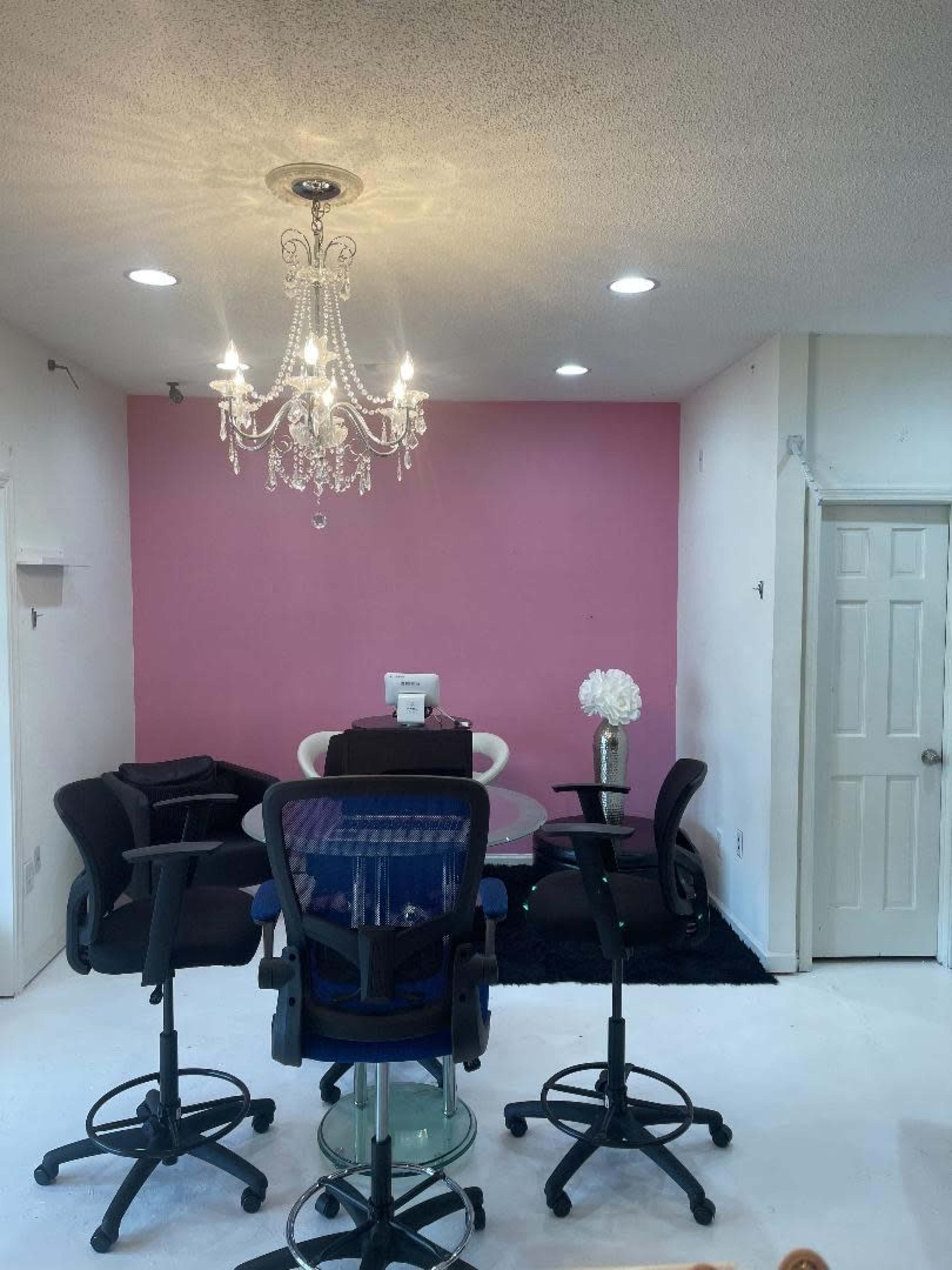 An office space with a glass table surrounded by several chairs, a chandelier overhead, and a pink accent wall.