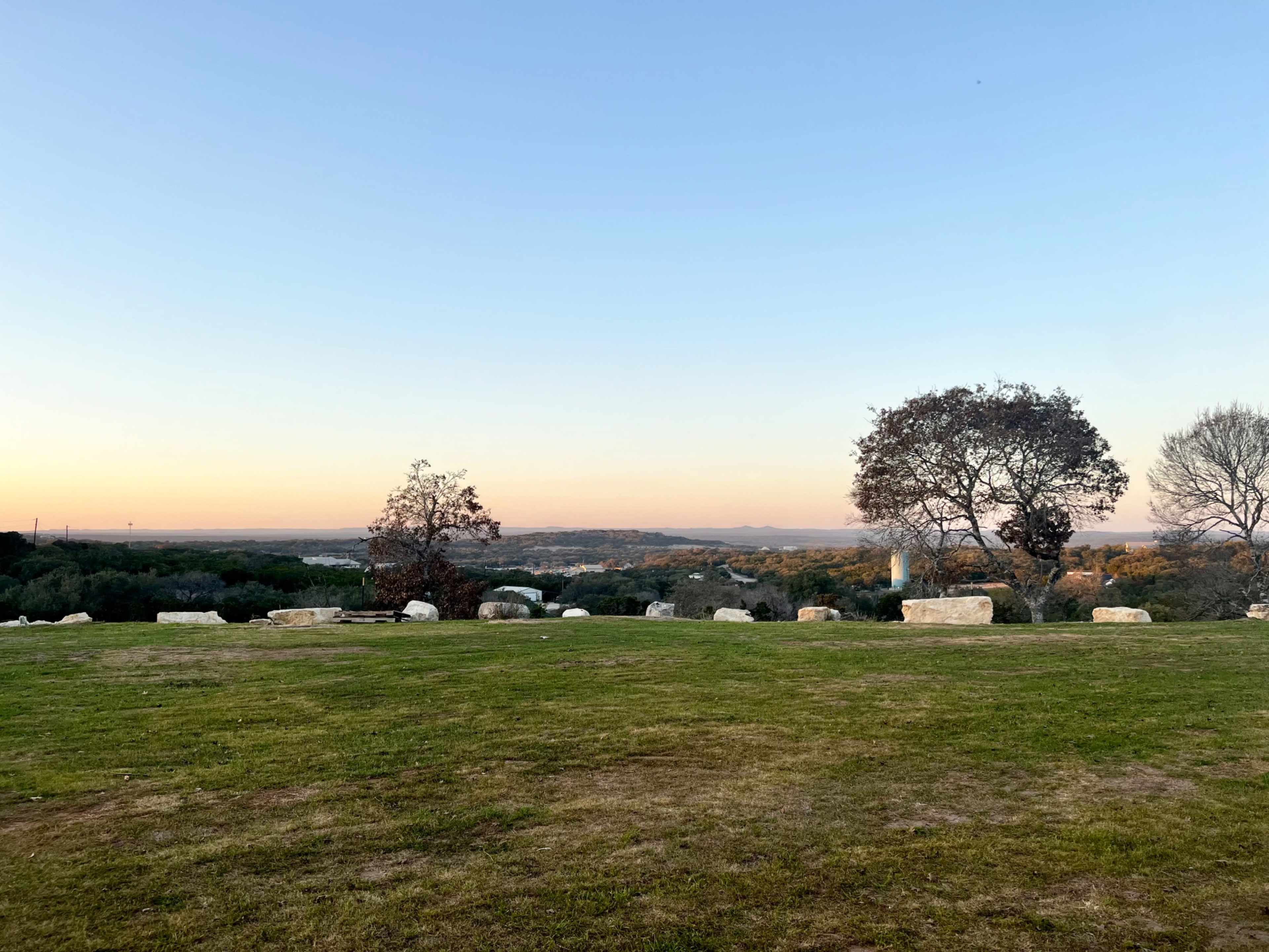 A grassy area with scattered stones, flanked by two trees, overlooking a valley at sunset.