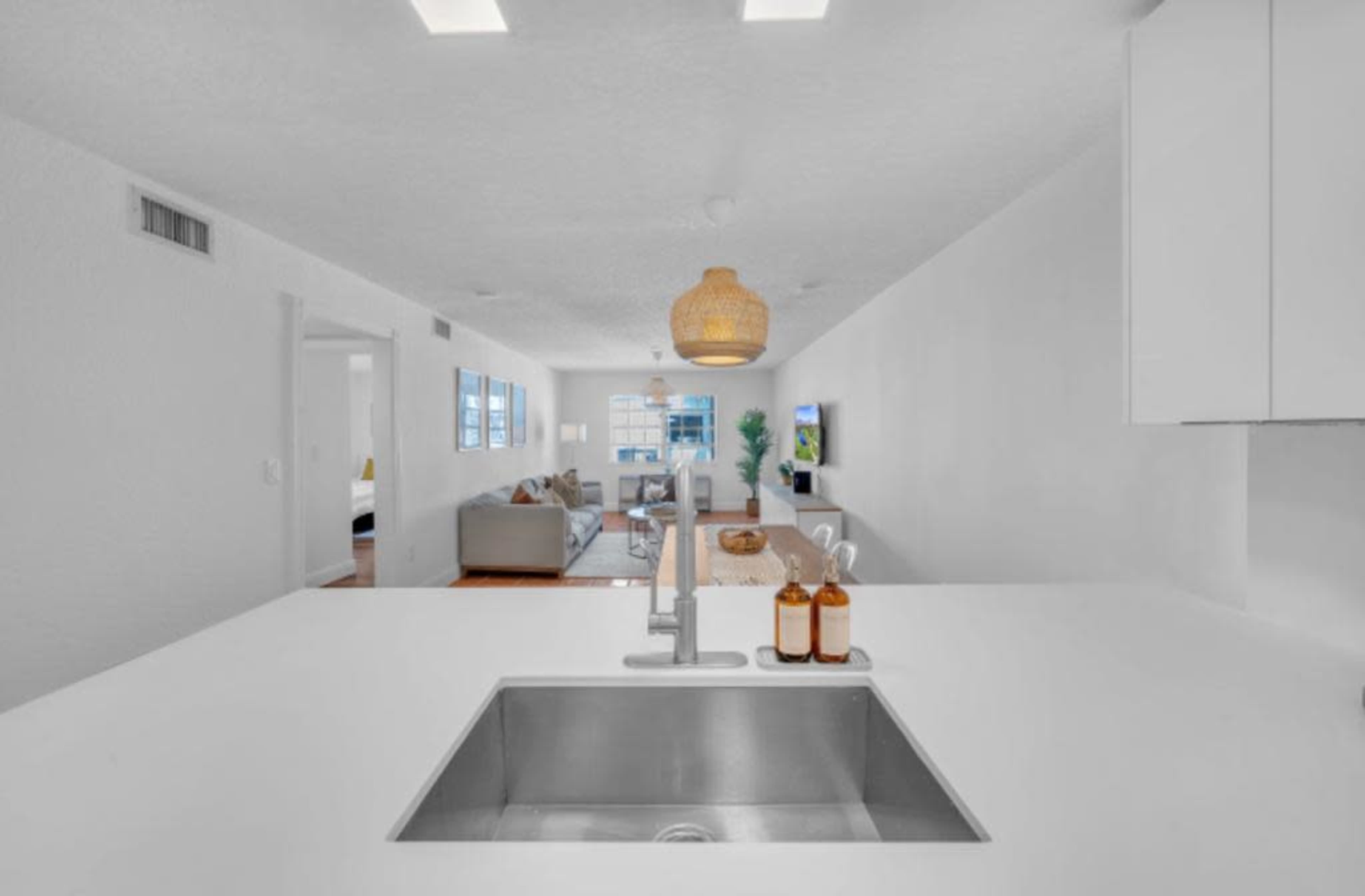 A modern kitchen features a stainless steel sink with decorative bottles and a view into a bright, open living area with a sofa and windows.