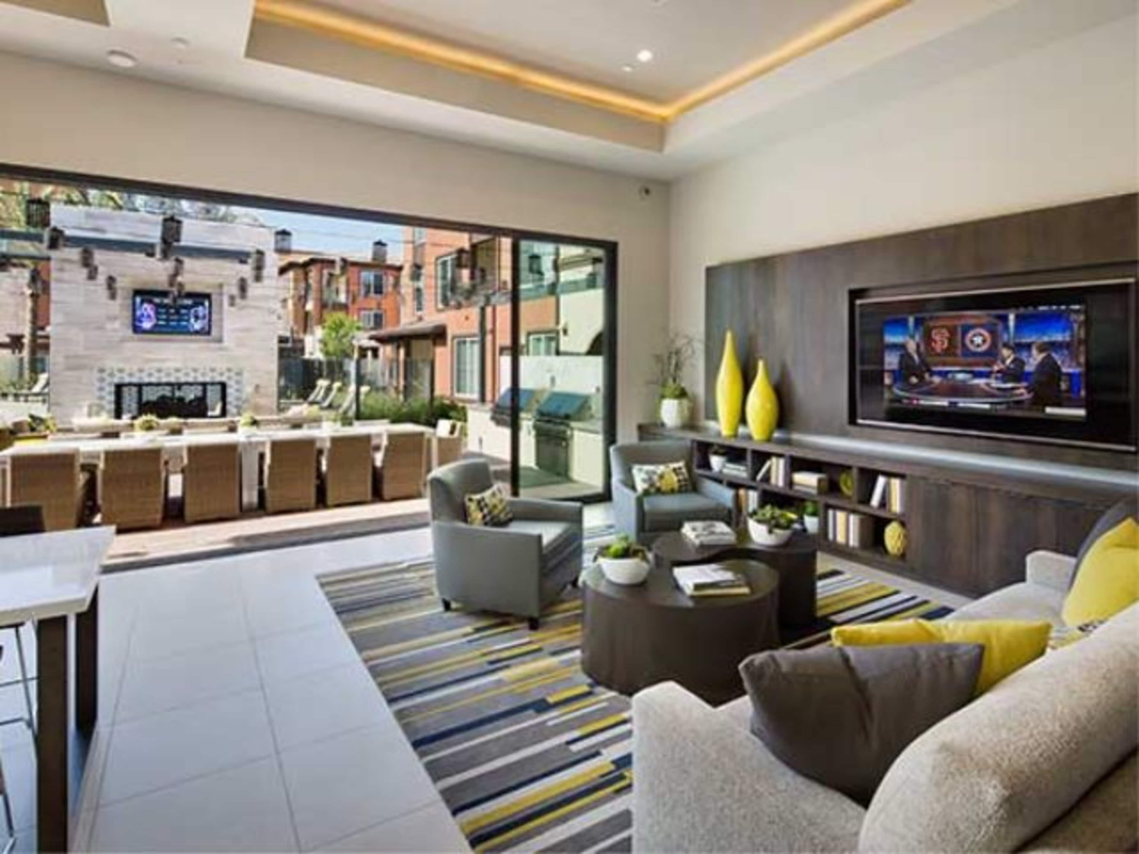 A modern living room features a large window showcasing an outdoor seating area, with a television mounted on the wall and a coffee table surrounded by gray chairs and a sofa.
