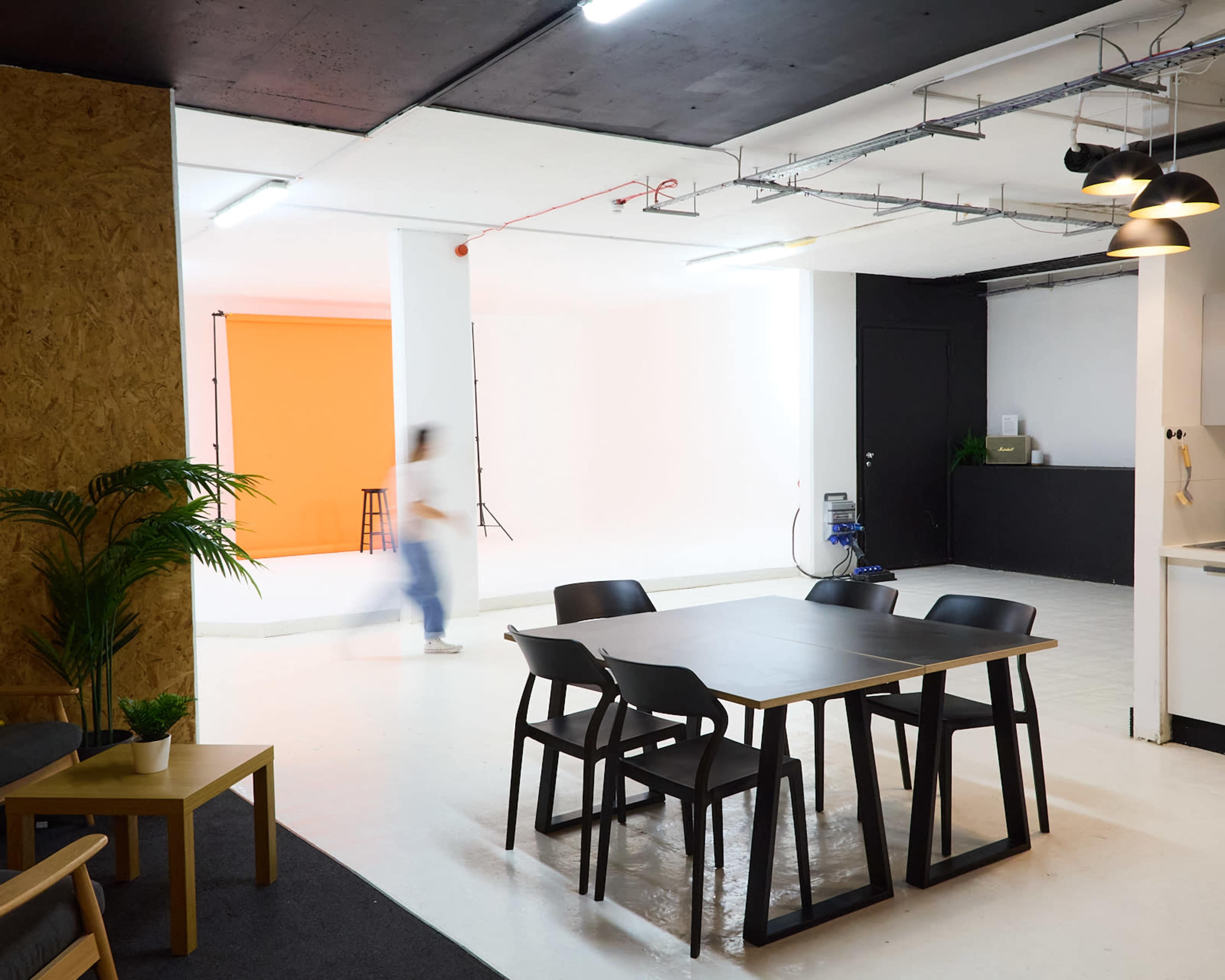A spacious studio featuring a dining table with chairs, an orange backdrop in one corner, and a white backdrop in another, with a blurred figure moving through the scene.