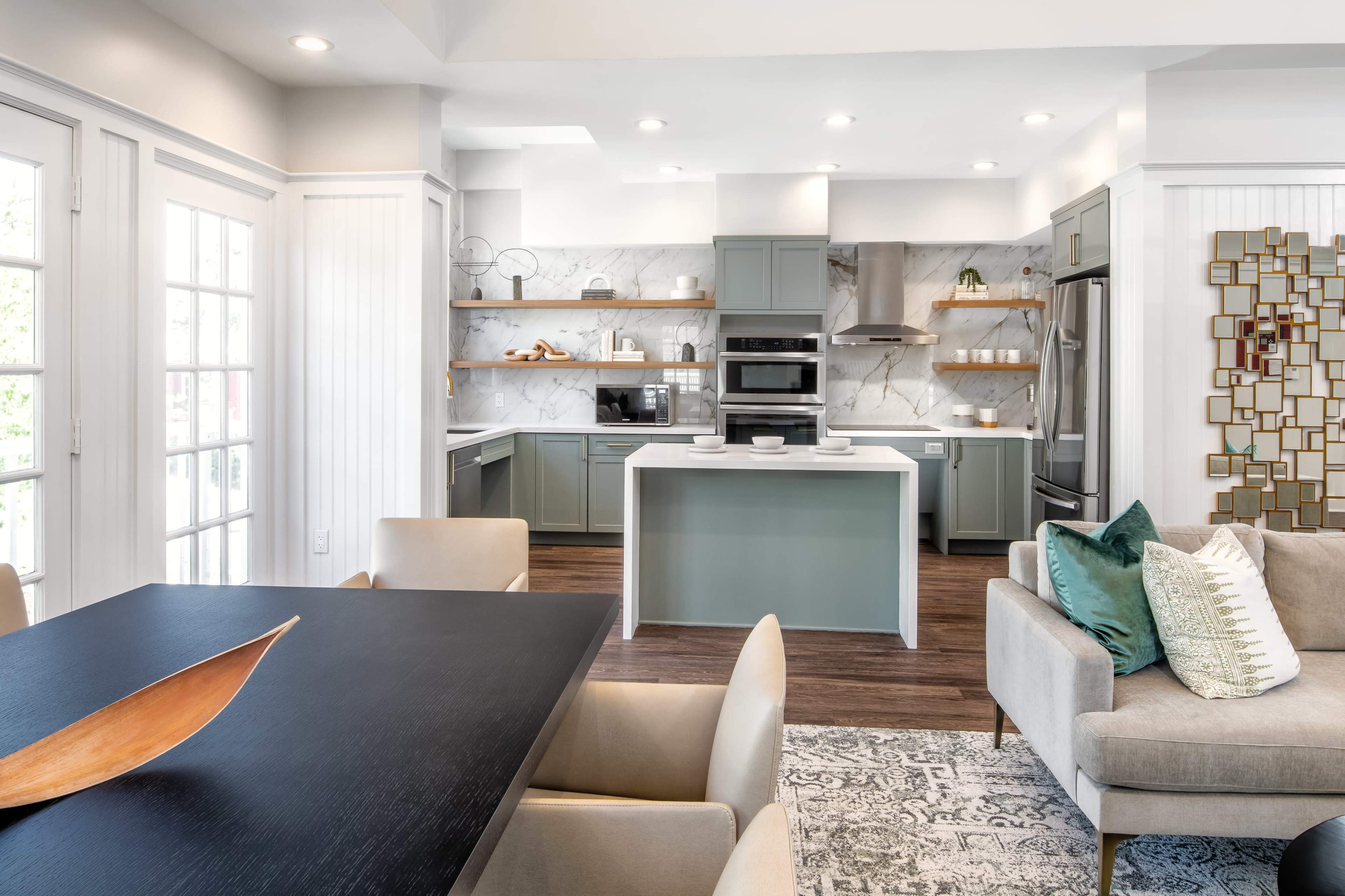 A modern kitchen with light green cabinets, stainless steel appliances, and a dining area featuring a dark wooden table and beige chairs.
