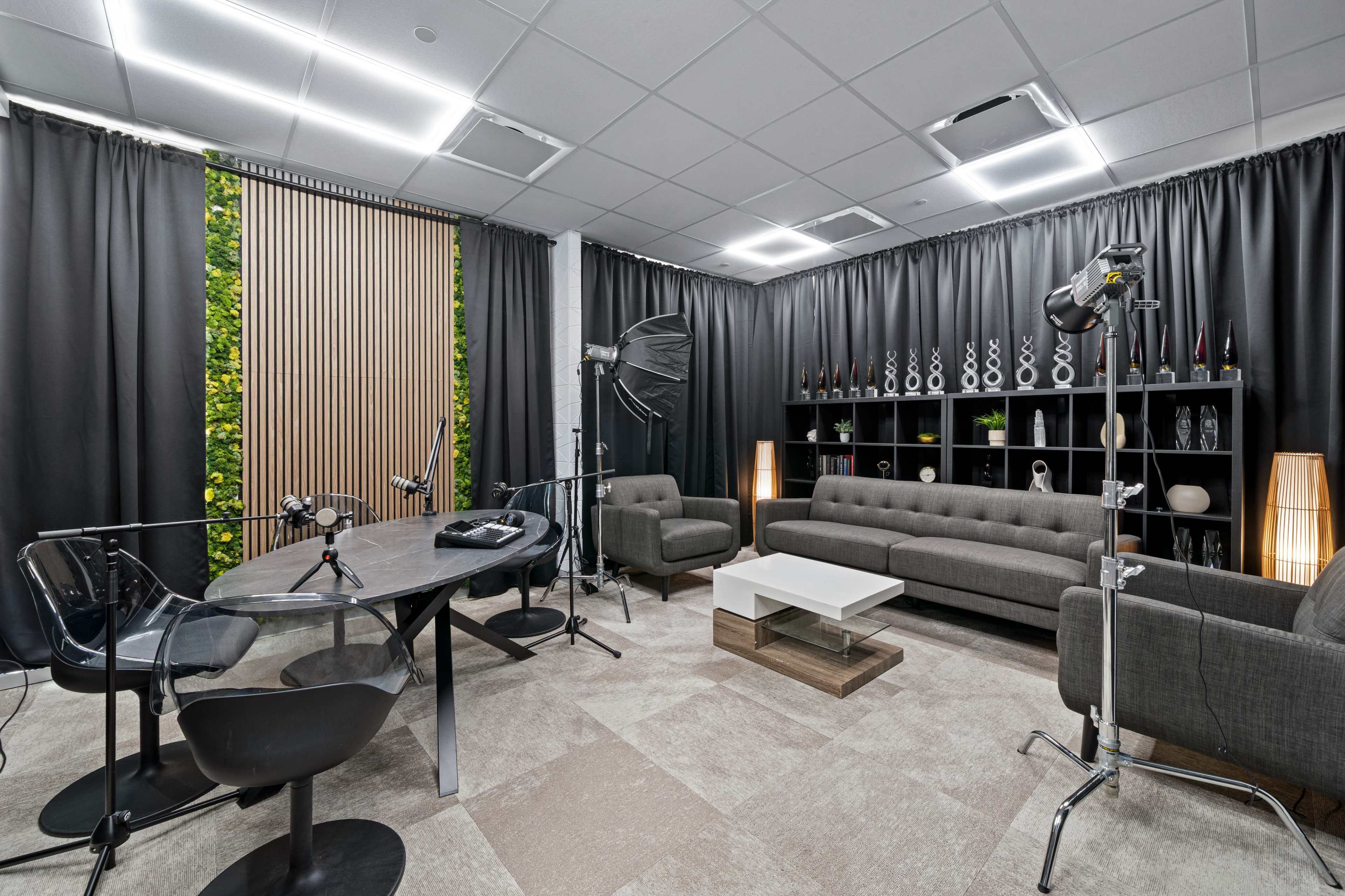 The image shows a modern studio space with a round table, gray sofas, lighting equipment, and a backdrop of greenery behind wooden slats.