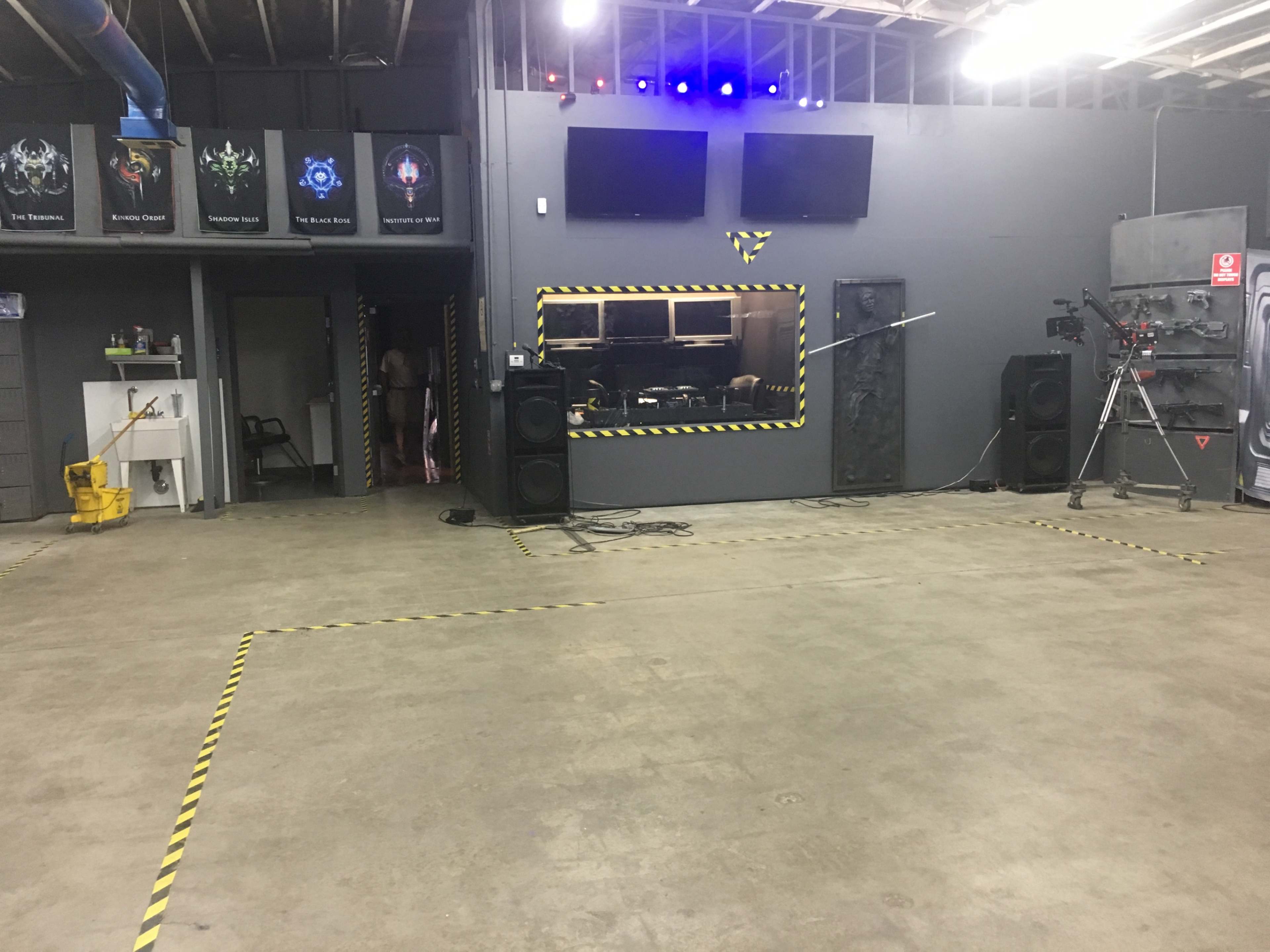 An empty indoor space with a concrete floor, sound equipment, a mirrored wall, and hazard-striped tape marking the perimeter.