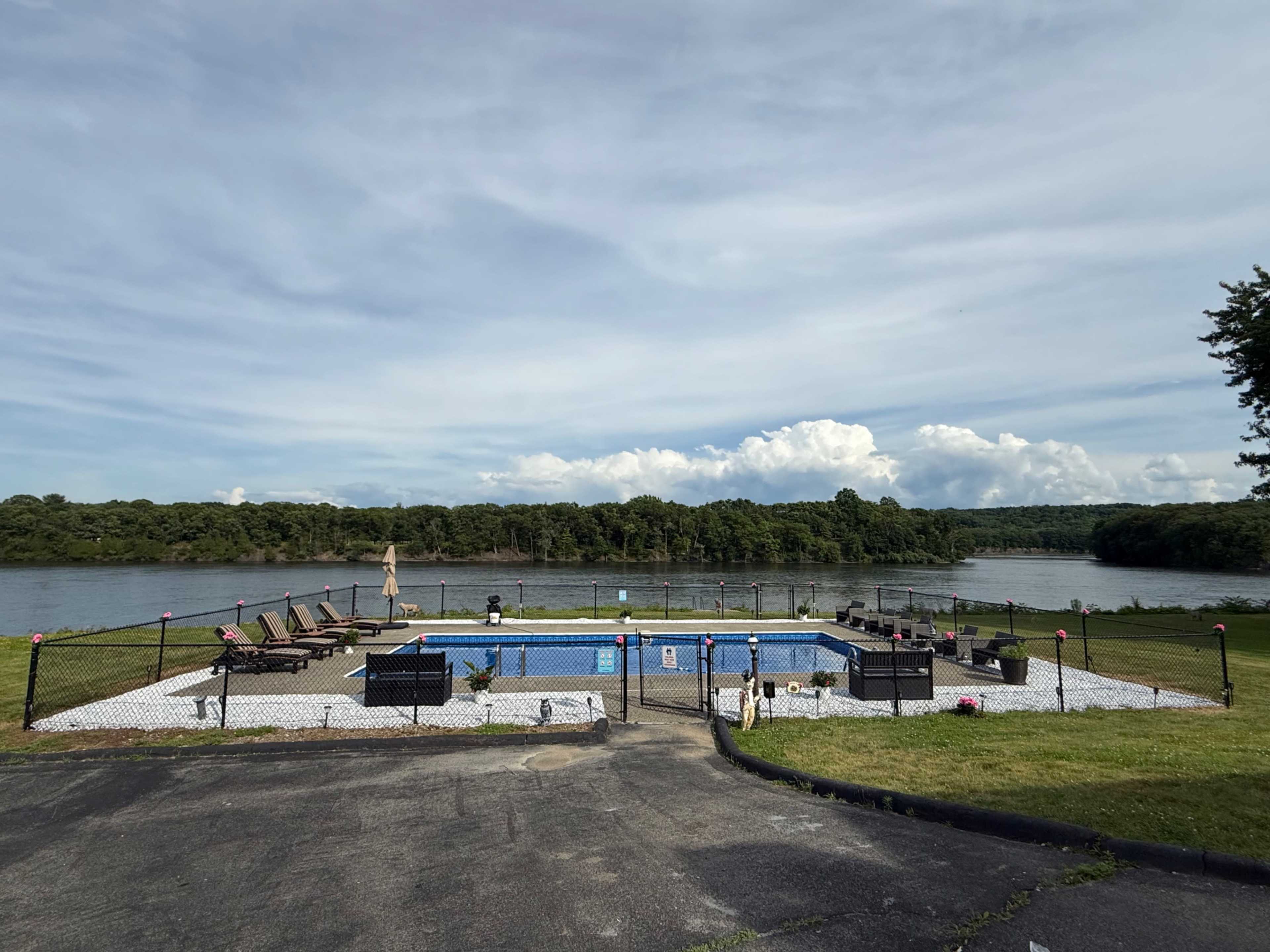 The image shows a fenced swimming pool area overlooking a calm body of water with green trees in the background.
