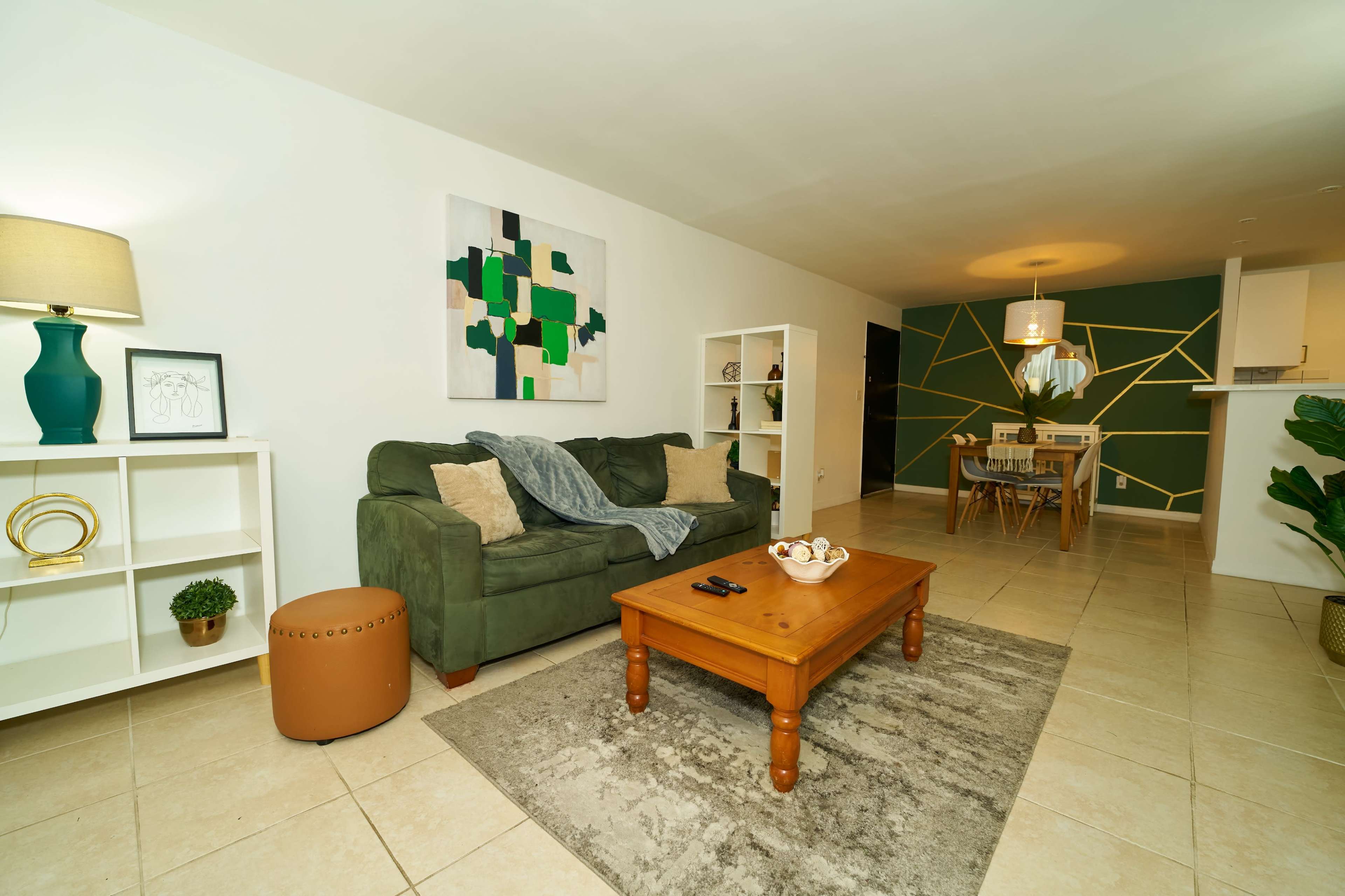 A living room features a green sofa, a wooden coffee table, and a dining area in the background with geometric wall decor.