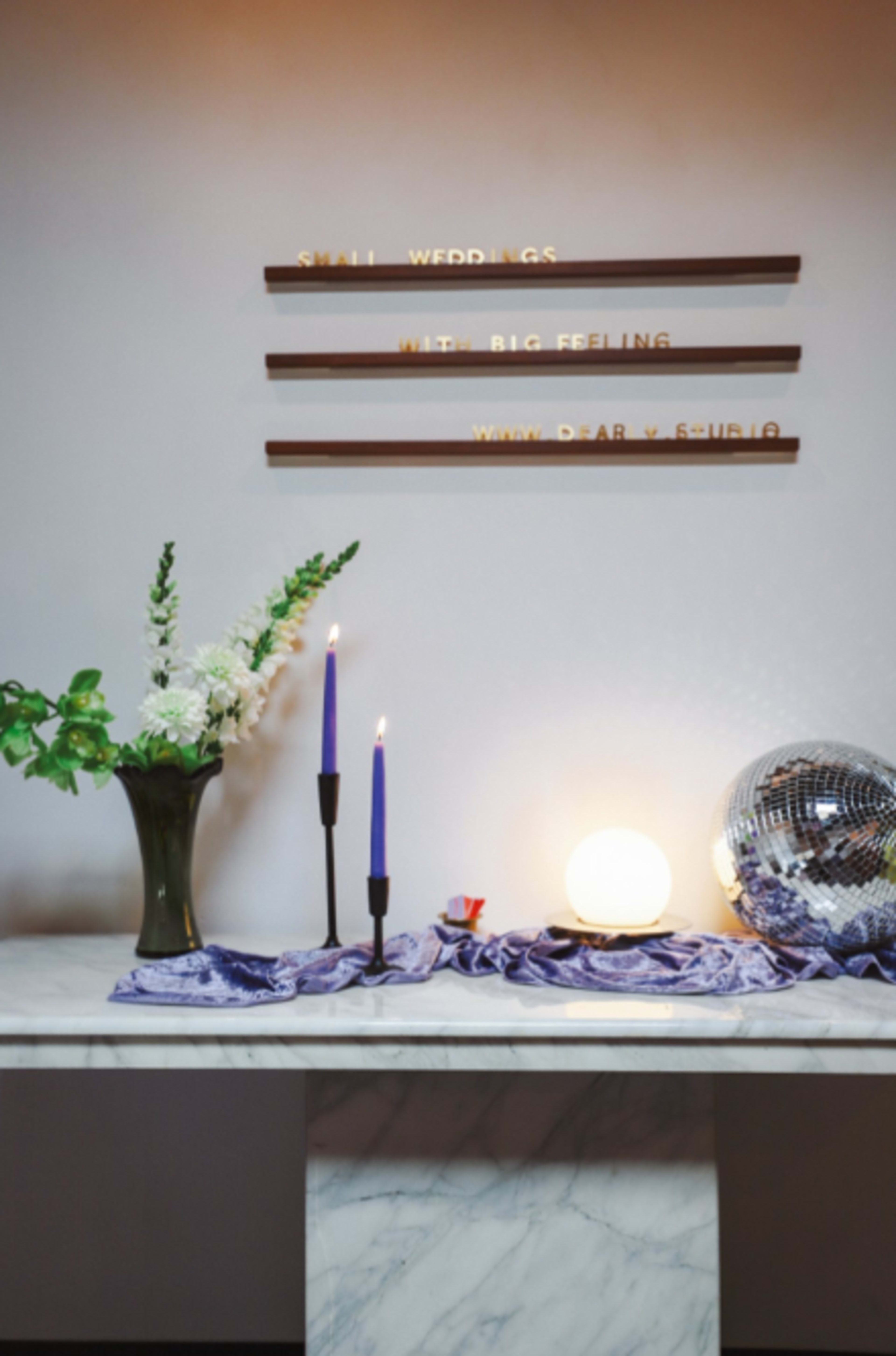 A marble table features two purple candles, a decorative vase of flowers, and a disco ball under a sign that reads "SMALL WEDDINGS. BIG FEELING."