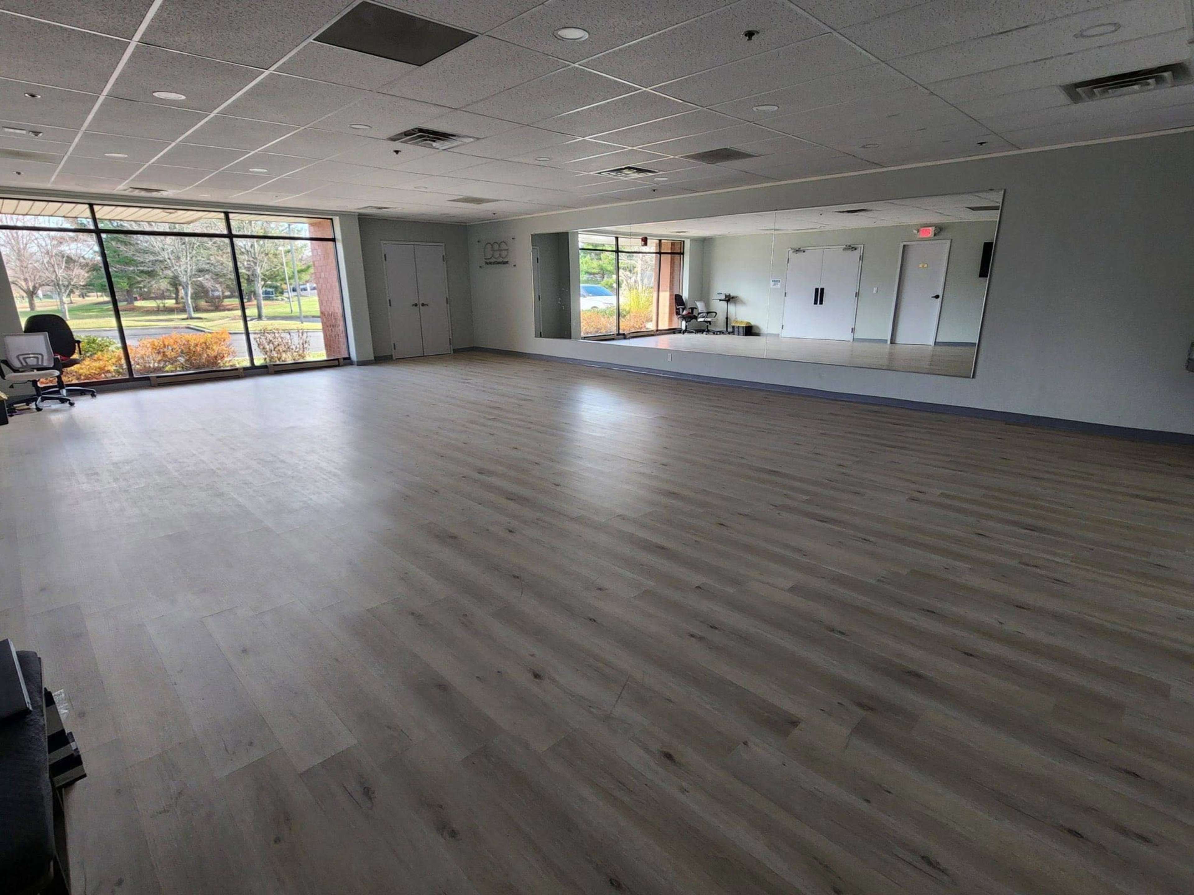 The image shows an empty, spacious room with large windows and a wall mirror, featuring light-colored wooden flooring.