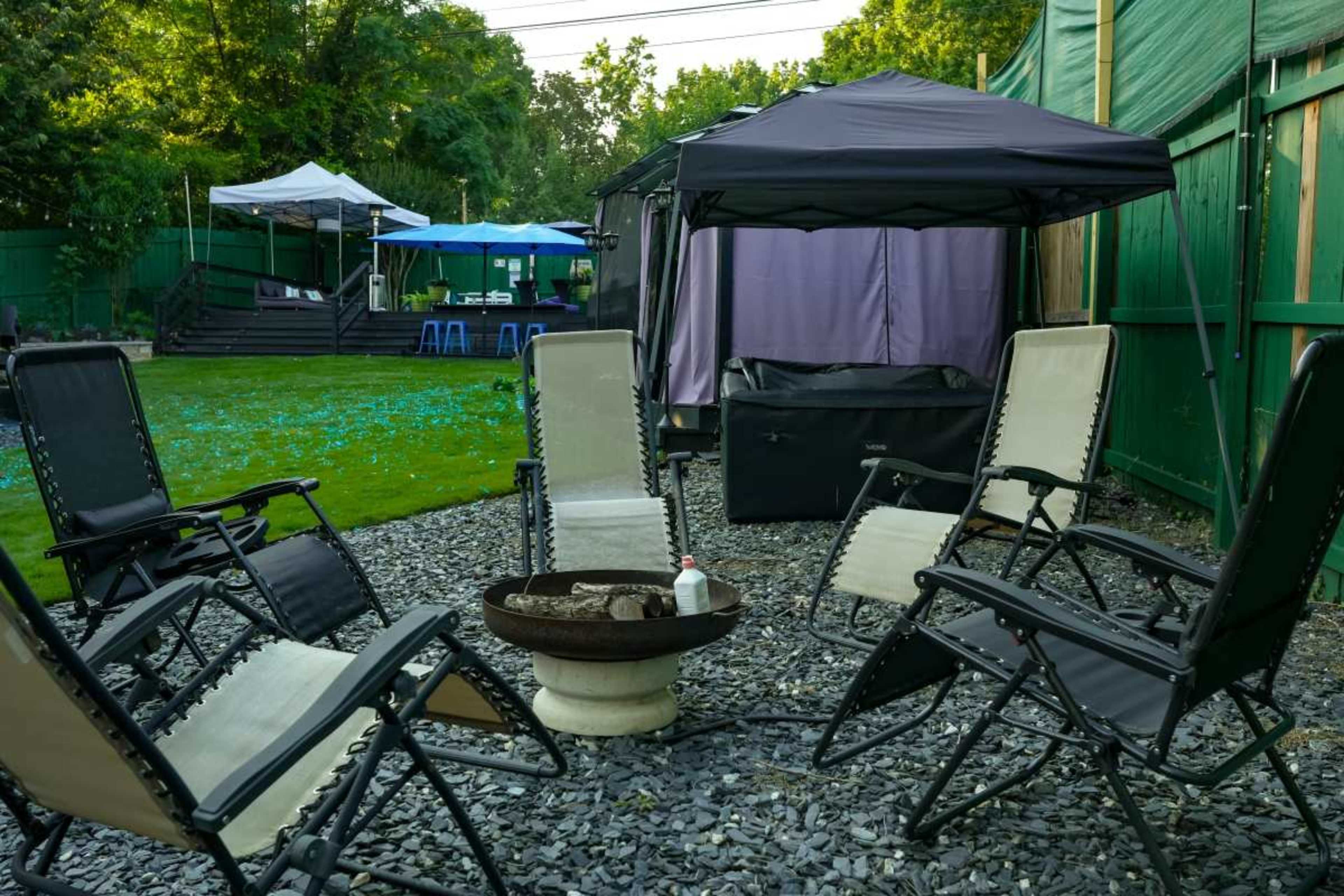The image shows a backyard setting with several folding chairs arranged around a fire pit on gravel, surrounded by green grass and tents in the background.