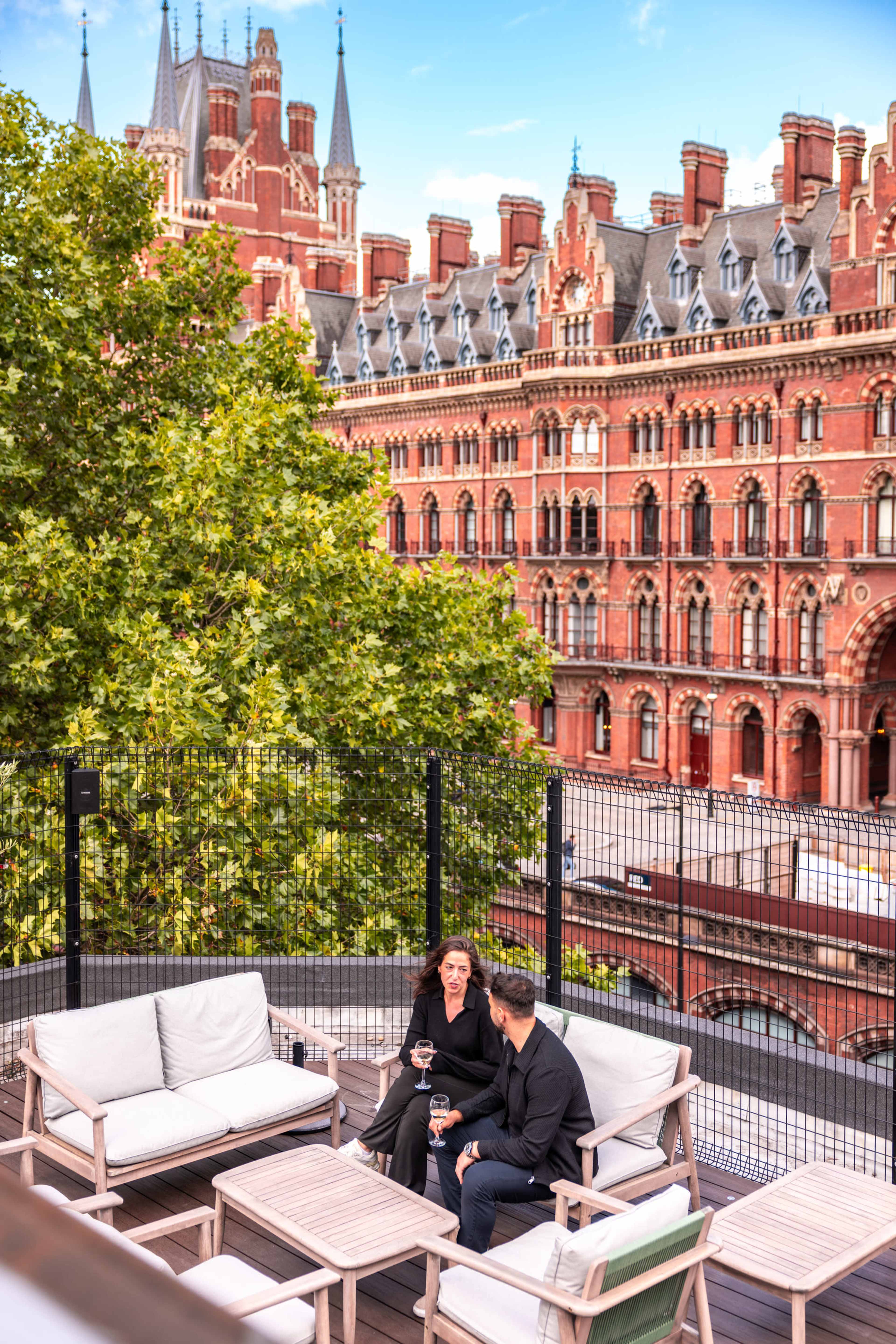 Two people sit on a terrace with stylish furniture, enjoying drinks while overlooking a historic building with red brick architecture.