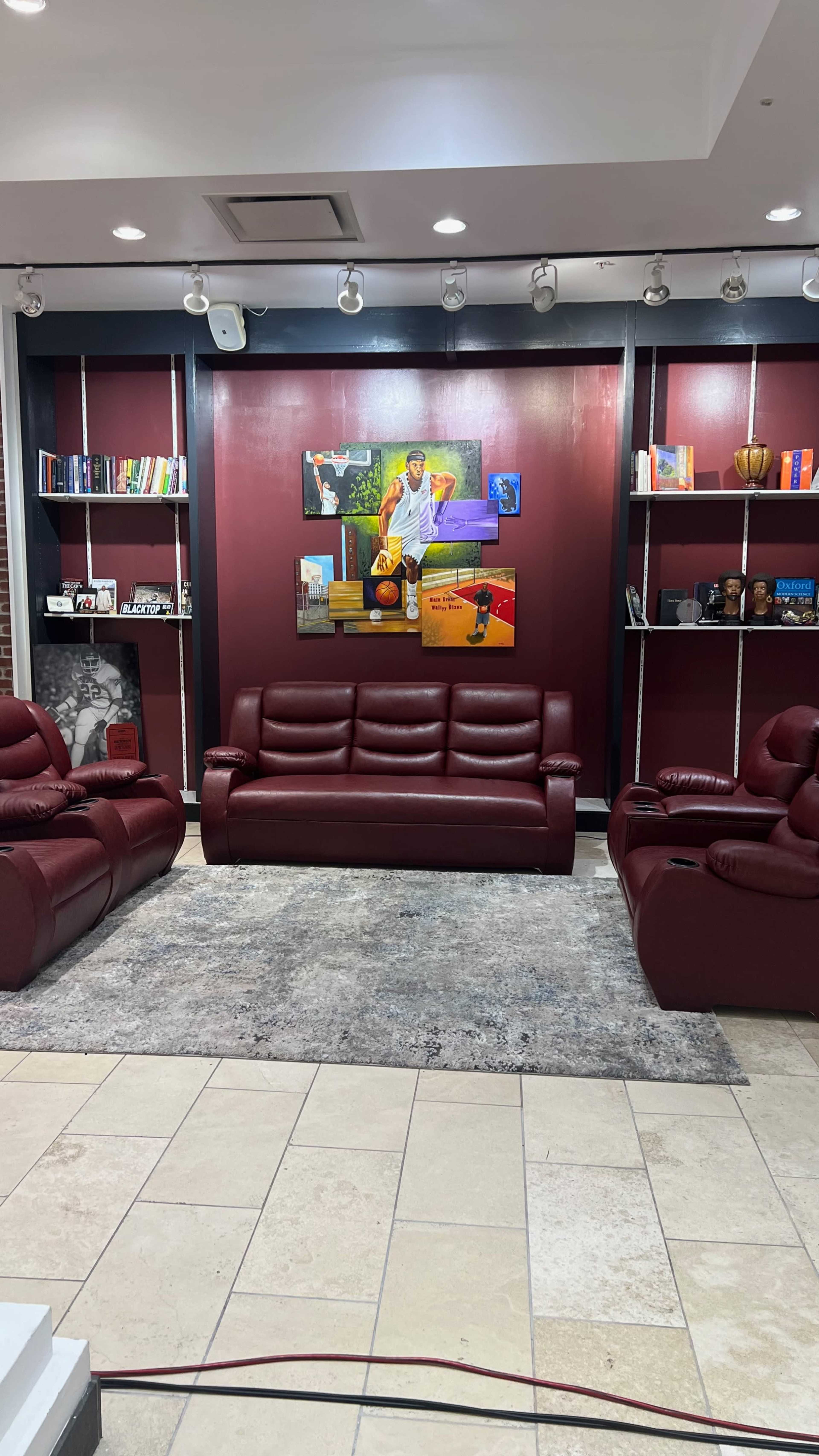 The image shows a modern lounge area with burgundy leather seating, a wall adorned with a colorful painting, and shelves displaying books and decorative items.