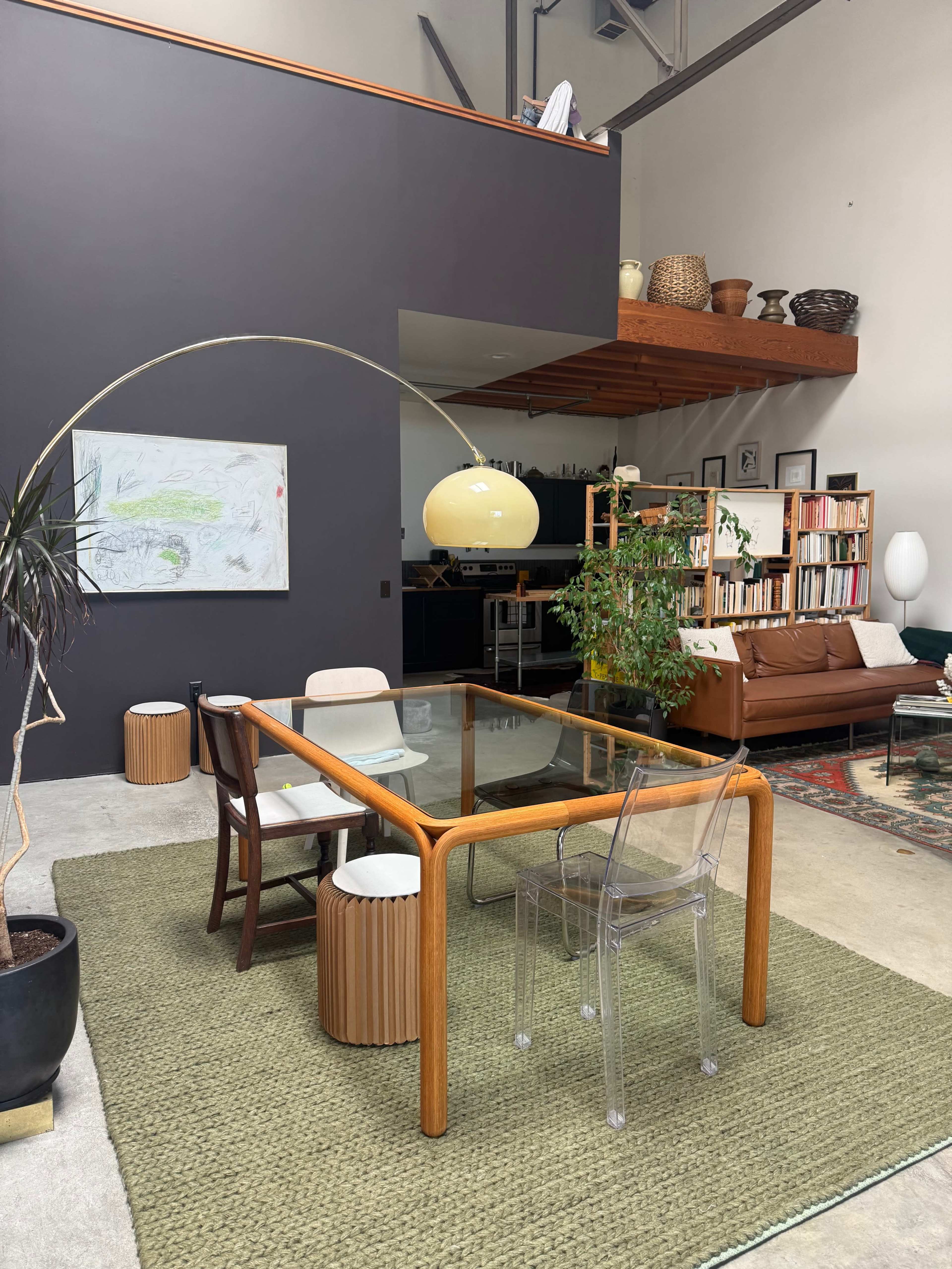 The image shows a modern dining area with a glass-topped wooden table, clear plastic chairs, a potted plant, and a curved floor lamp, all set against a backdrop of bookshelves and a leather sofa.