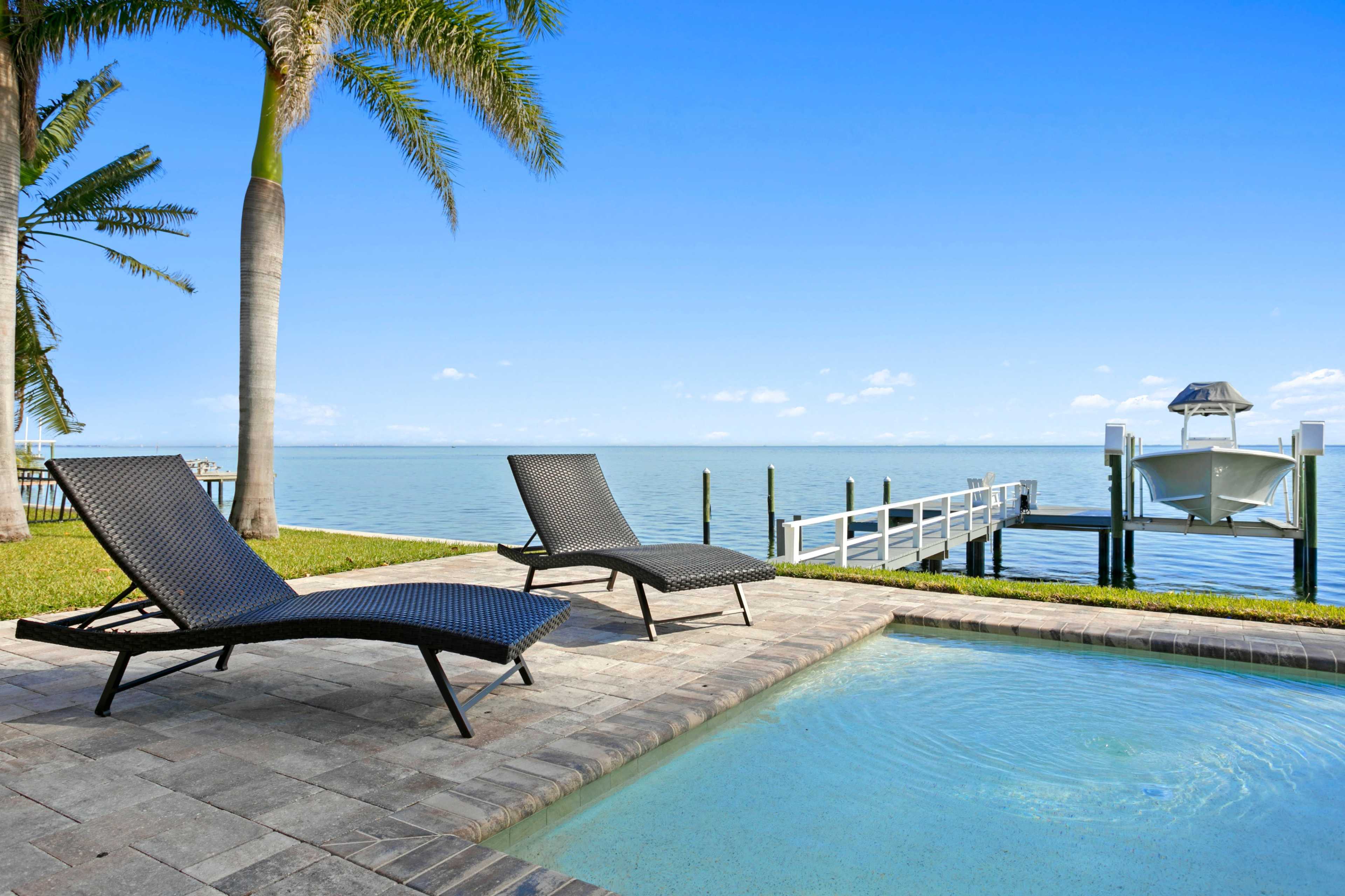 The image shows two lounge chairs beside a swimming pool overlooking a calm body of water and a dock.