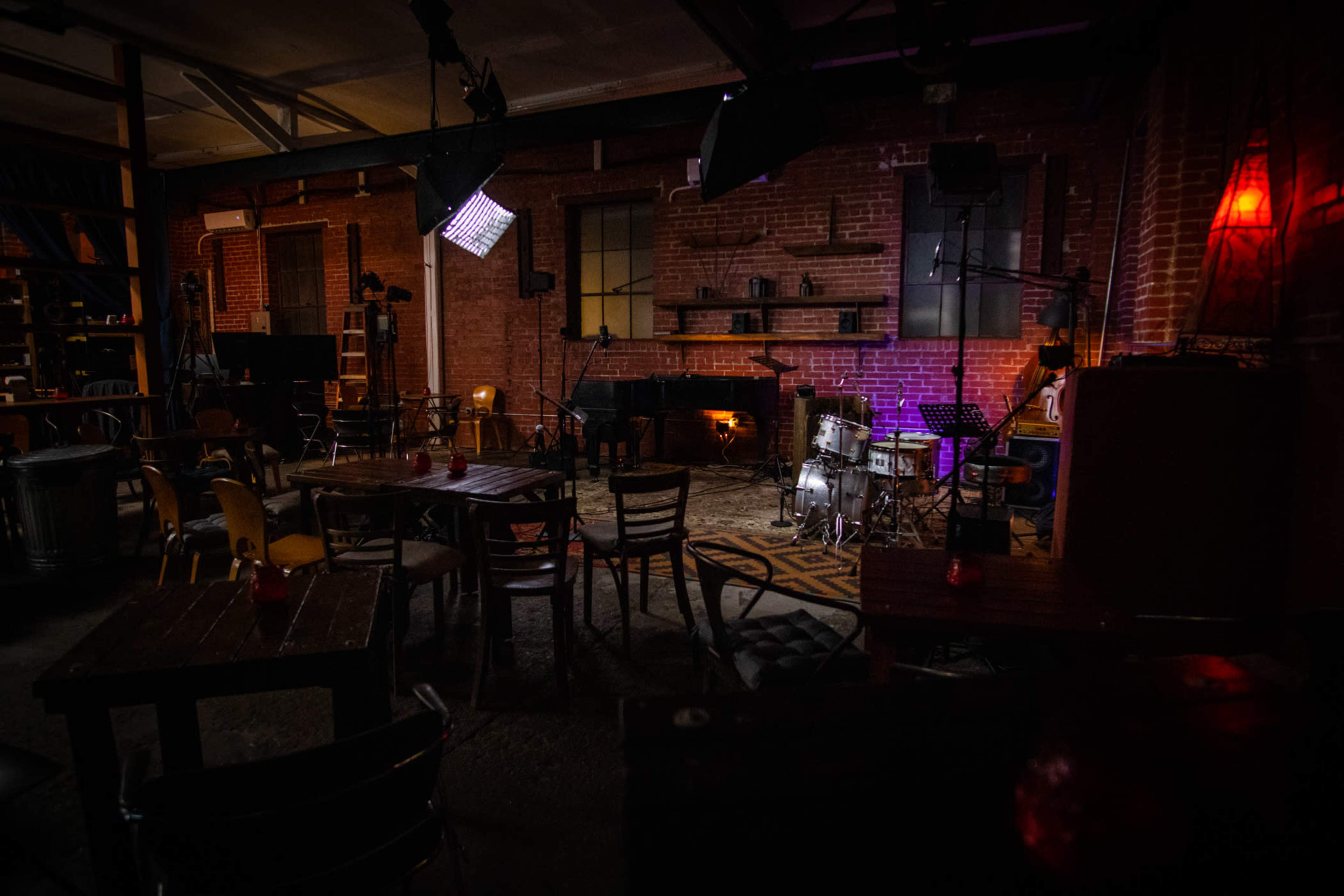 A music venue with a drum set and stage equipment, surrounded by wooden tables and chairs in a brick-walled room.