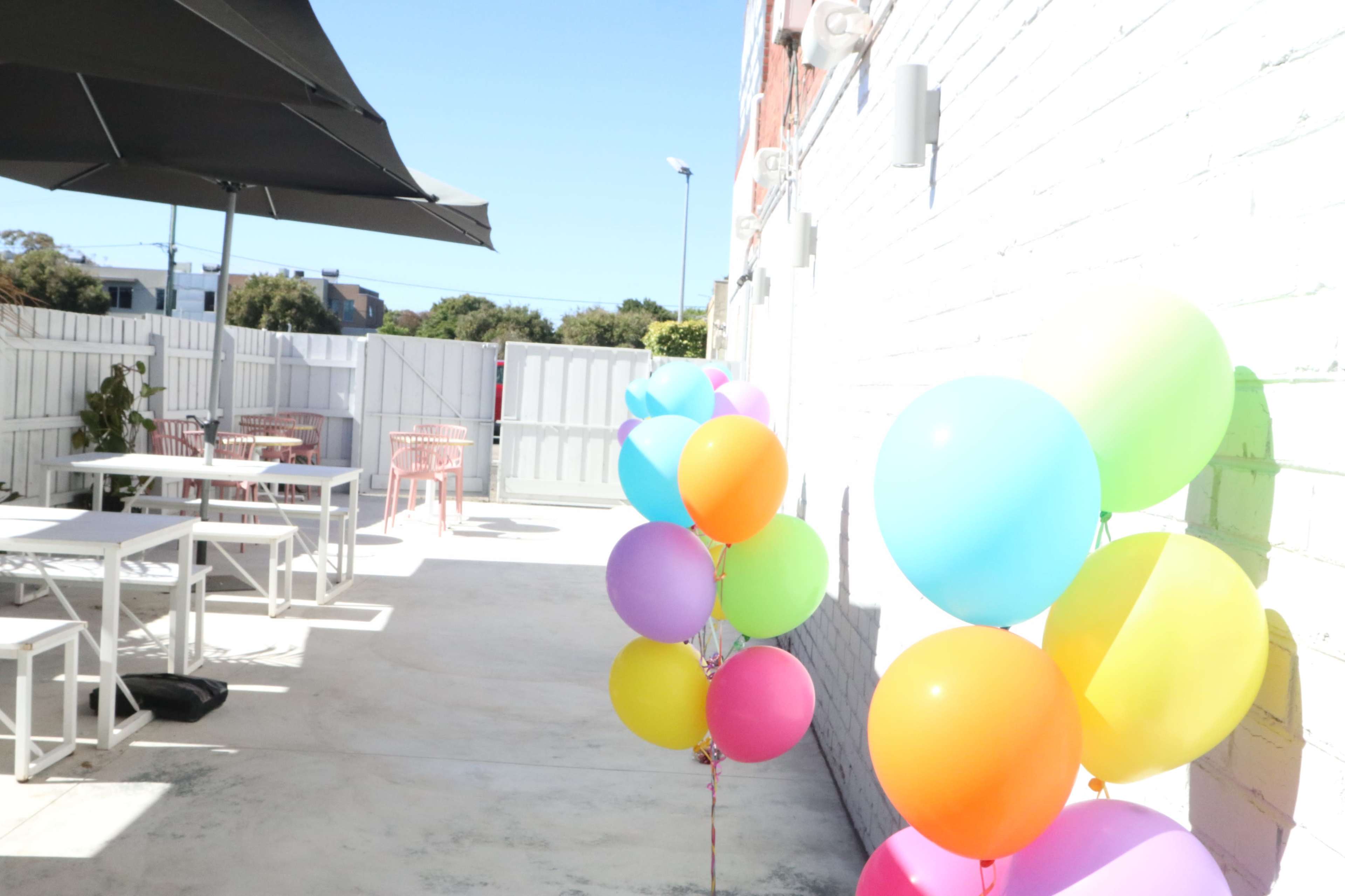 A sunny outdoor space with colorful balloons and white furniture, featuring a shaded area with an umbrella.